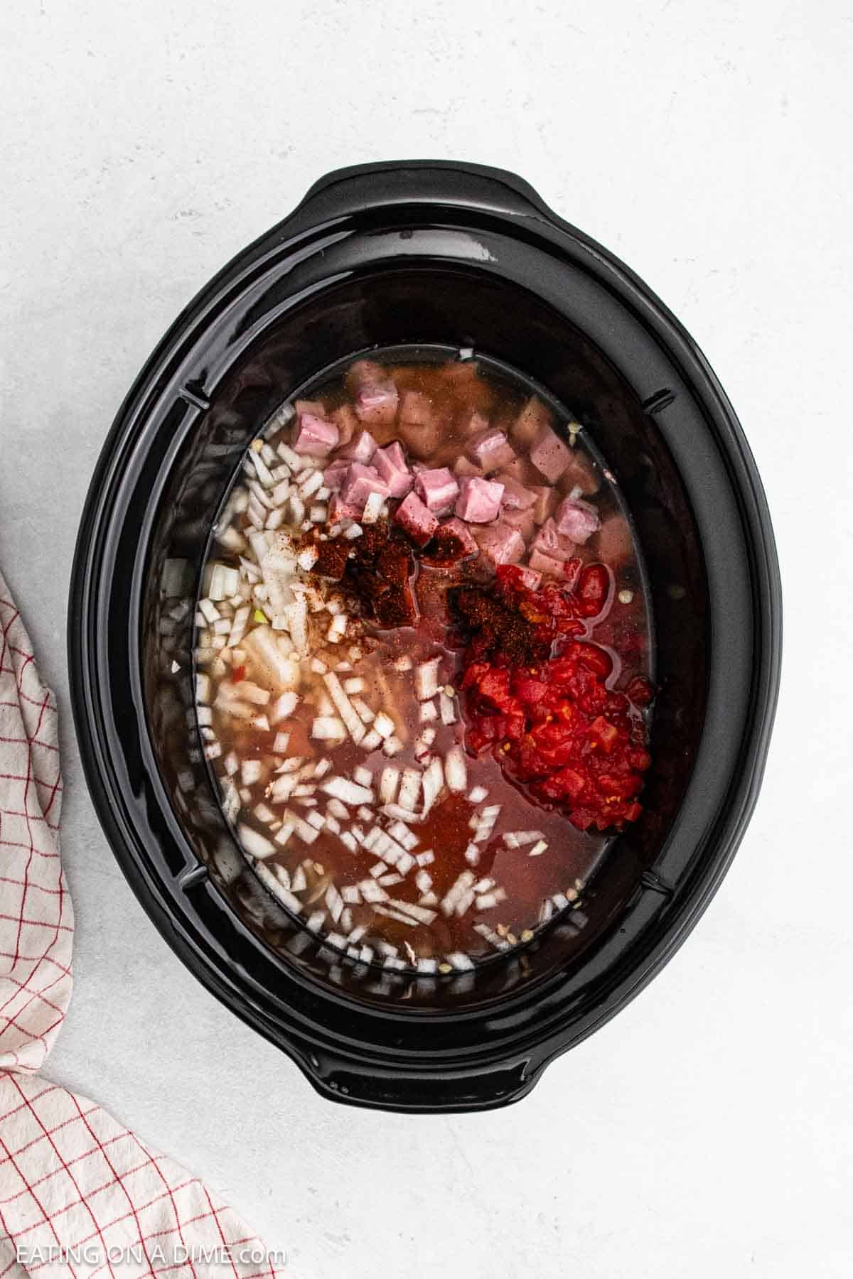 A crock pot brimming with diced onions, chopped tomatoes, cubed ham, and sliced red peppers swimming in a savory broth. The scene is reminiscent of a cozy kitchen preparing 15 bean soup with ham, with a red checkered cloth peeking in on the left side.