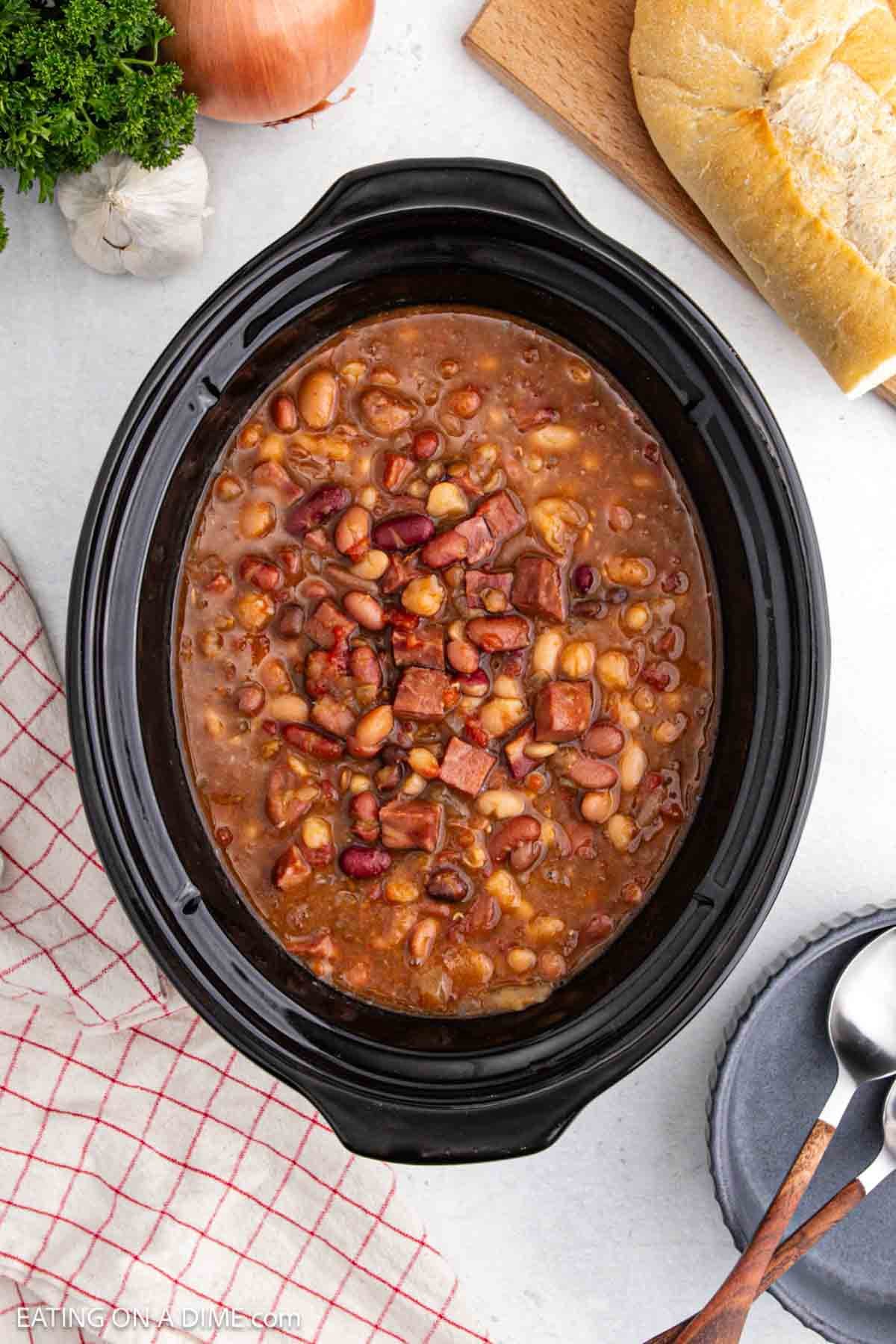 A crockpot brimming with crock pot 15 bean soup with ham, featuring tender chunks of meat and assorted beans. Beside the simmering pot are a rustic loaf of bread, a wooden spoon, a checked cloth, and a bowl with a spoon, all atop a light countertop.