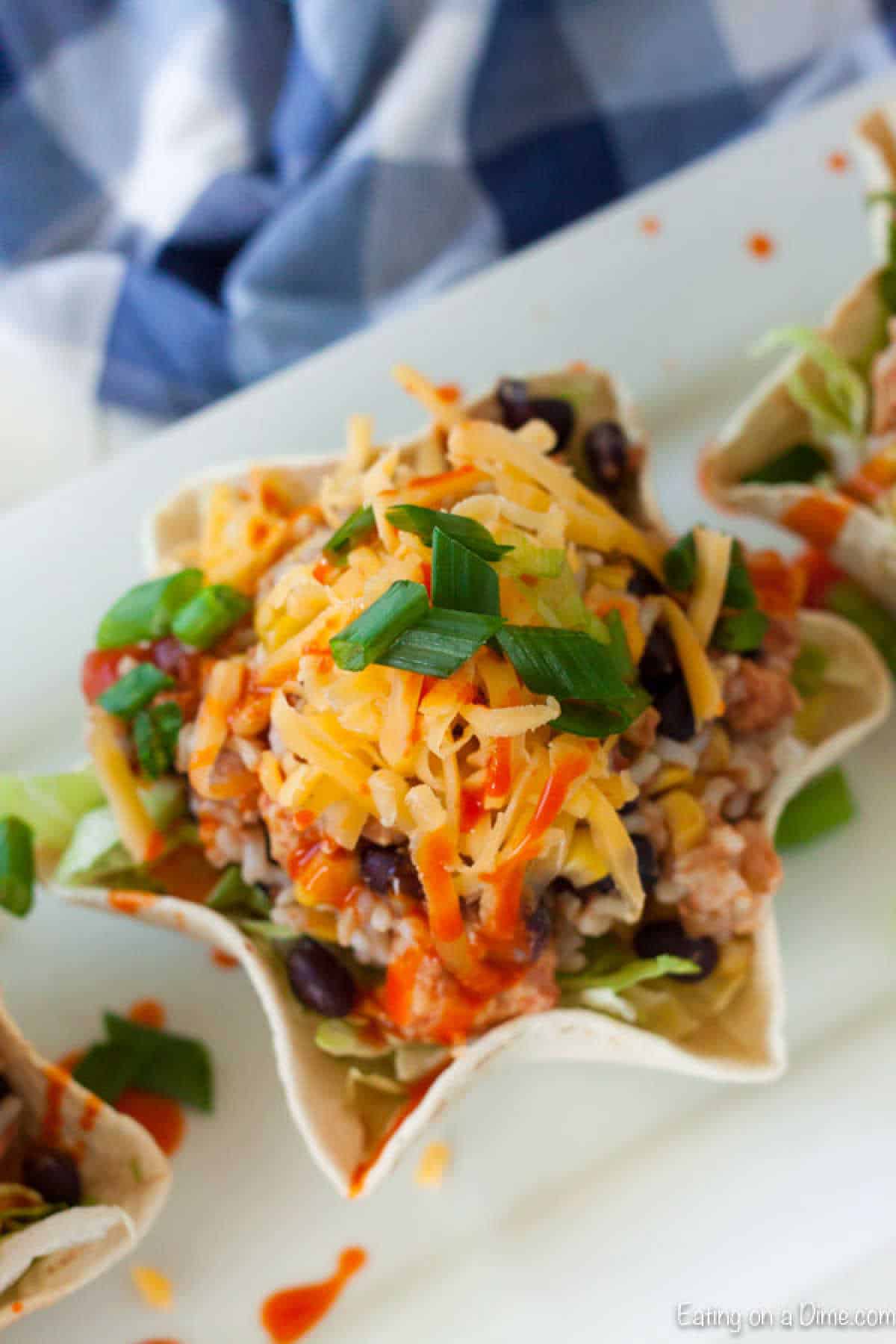 A taco salad in a tortilla bowl with shredded cheese, black beans, chopped green onions, and hot sauce drizzle brings to mind our Slow Cooker Chicken Burrito Bowl Recipe. A blue and white checkered cloth is casually draped in the background.