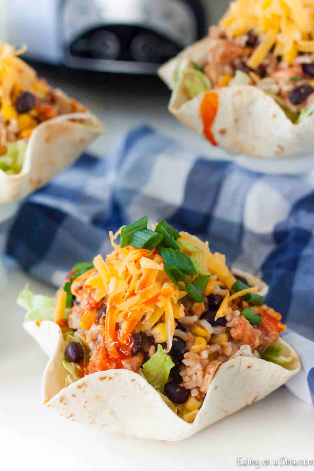 Taco bowls filled with rice, black beans, corn, lettuce, and diced tomatoes, topped with cheese and green onions. Crafted from tortillas, these bowls complement any Slow Cooker Chicken Burrito Bowl Recipe. A checkered blue cloth adds a touch of charm in the background.