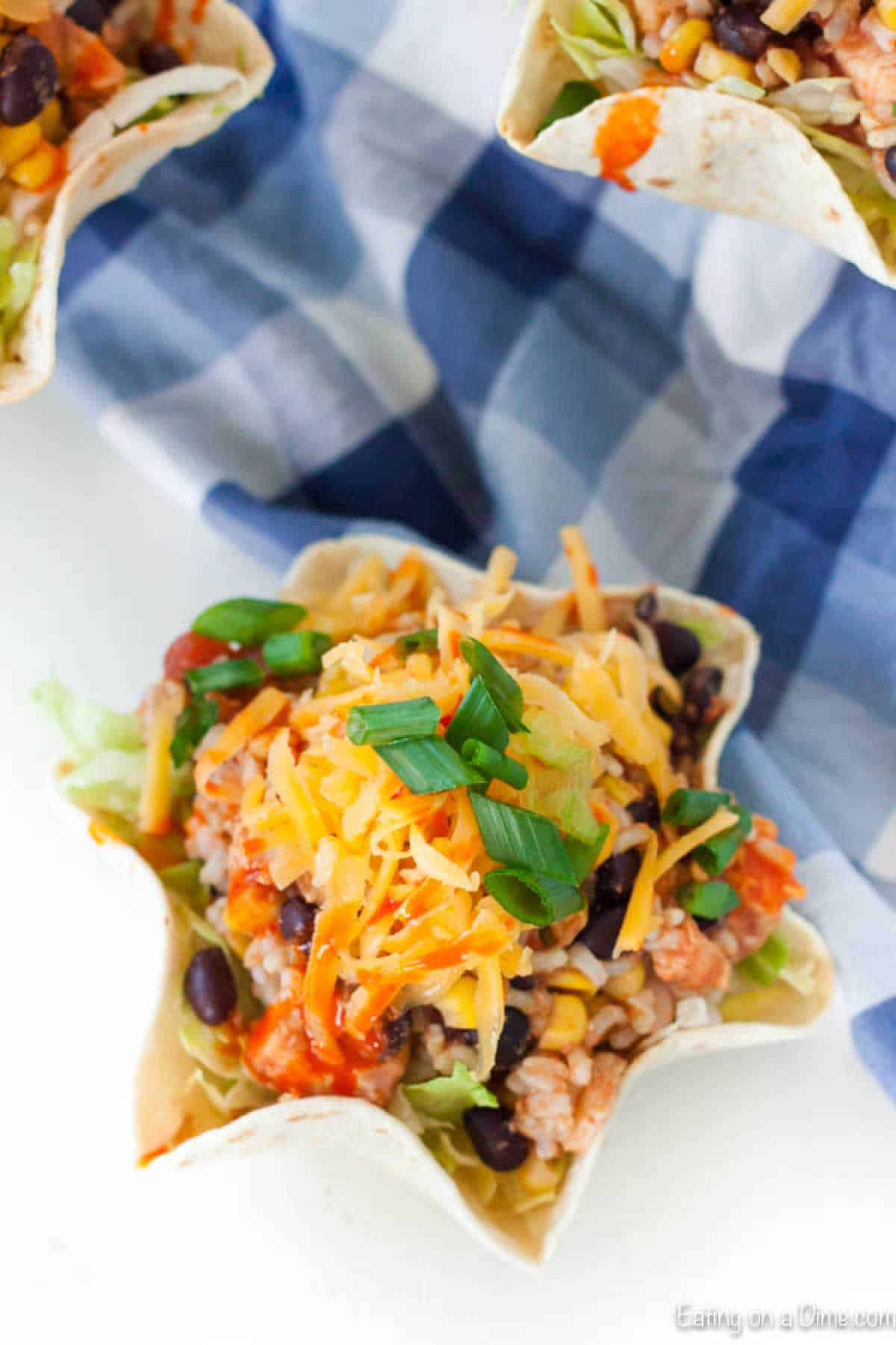 A taco salad with shredded cheese, black beans, corn, lettuce, and green onions served in a tortilla bowl. Inspired by a Slow Cooker Chicken Burrito Bowl Recipe, this dish rests on a white surface with a blue and white checkered cloth.