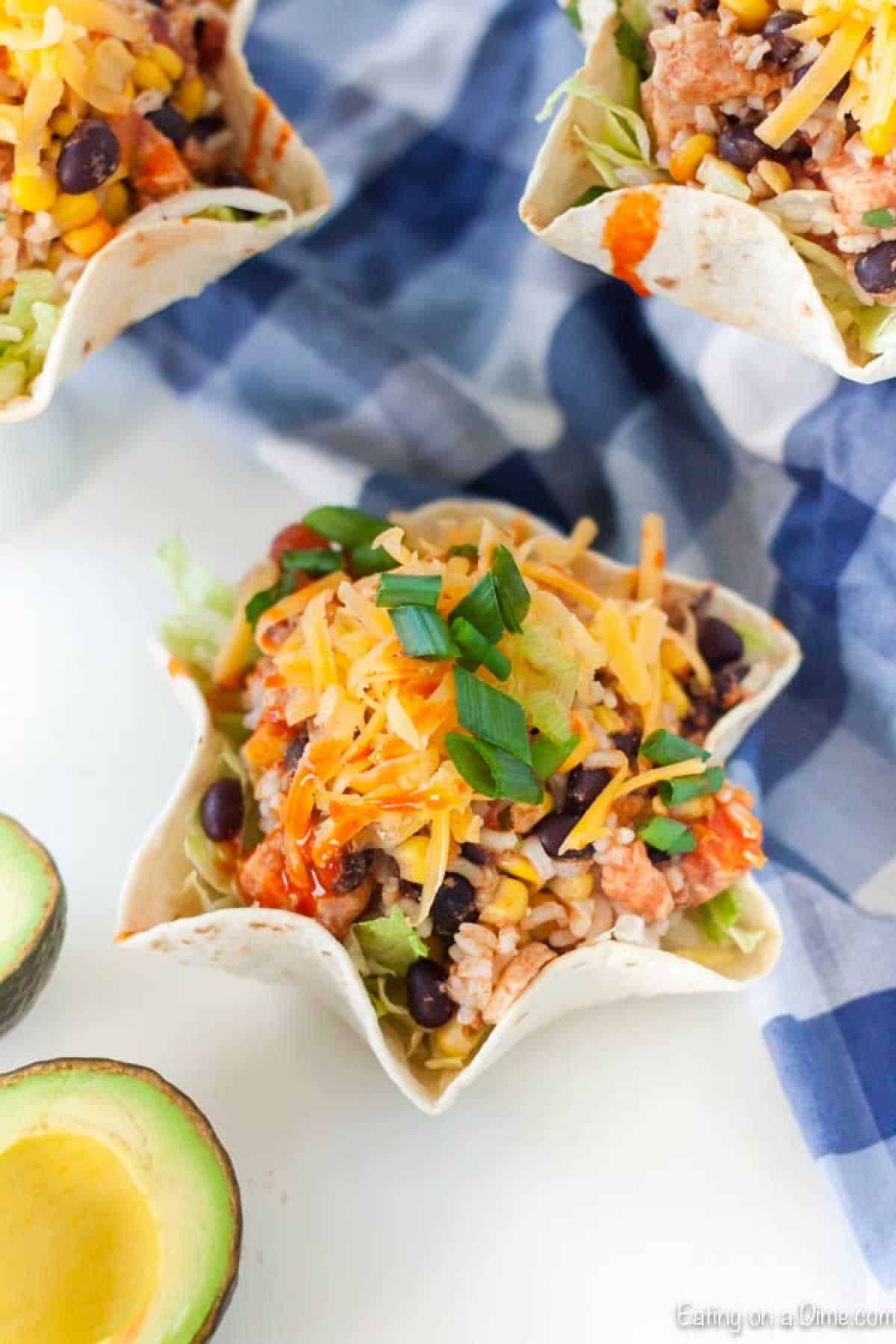 Tortilla bowls brimming with mixed salad, black beans, corn, shredded cheese, and green onions rest on a checkered cloth, embodying the essence of a slow cooker chicken burrito bowl recipe. A ripe avocado graces the scene in the foreground.