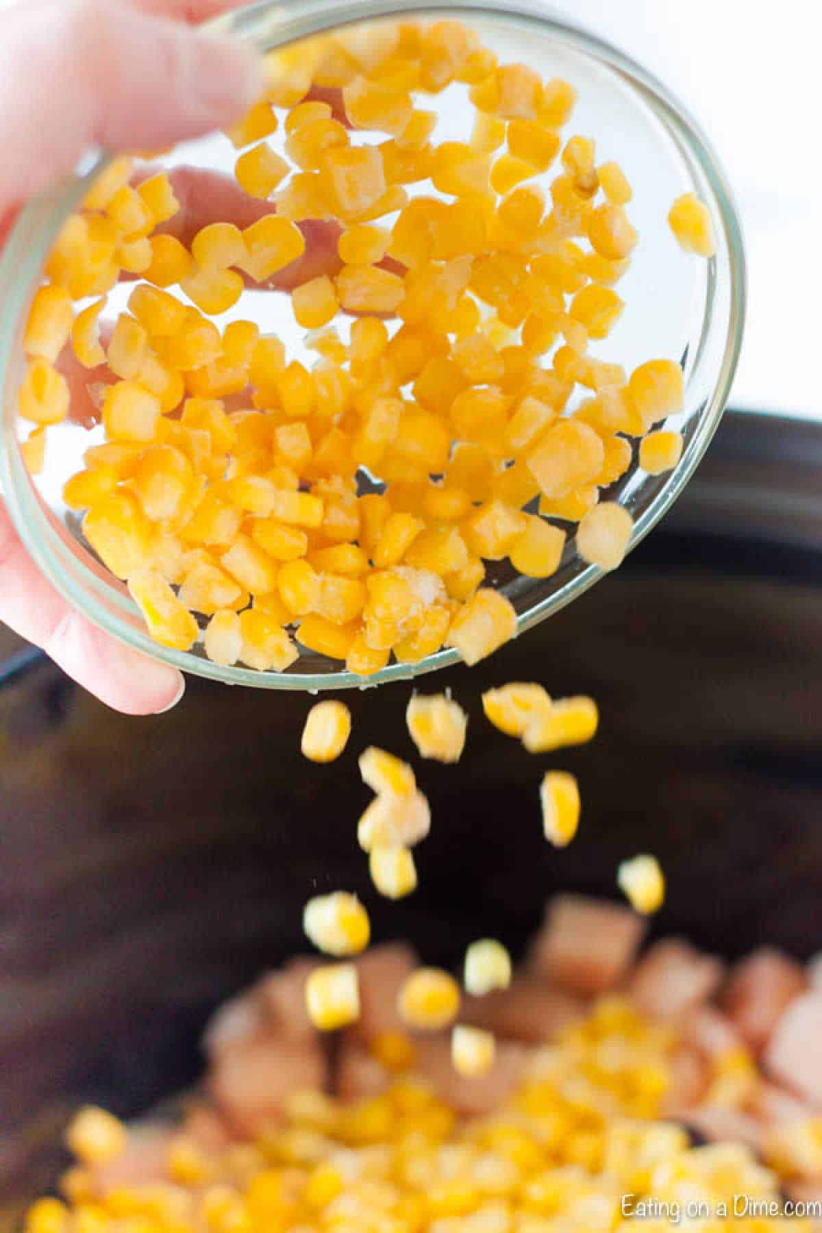 A hand is pouring a bowl of frozen corn kernels into a slow cooker brimming with diced vegetables, hinting at the start of a delicious Slow Cooker Chicken Burrito Bowl recipe. The image teases the promise of a hearty meal in the making.