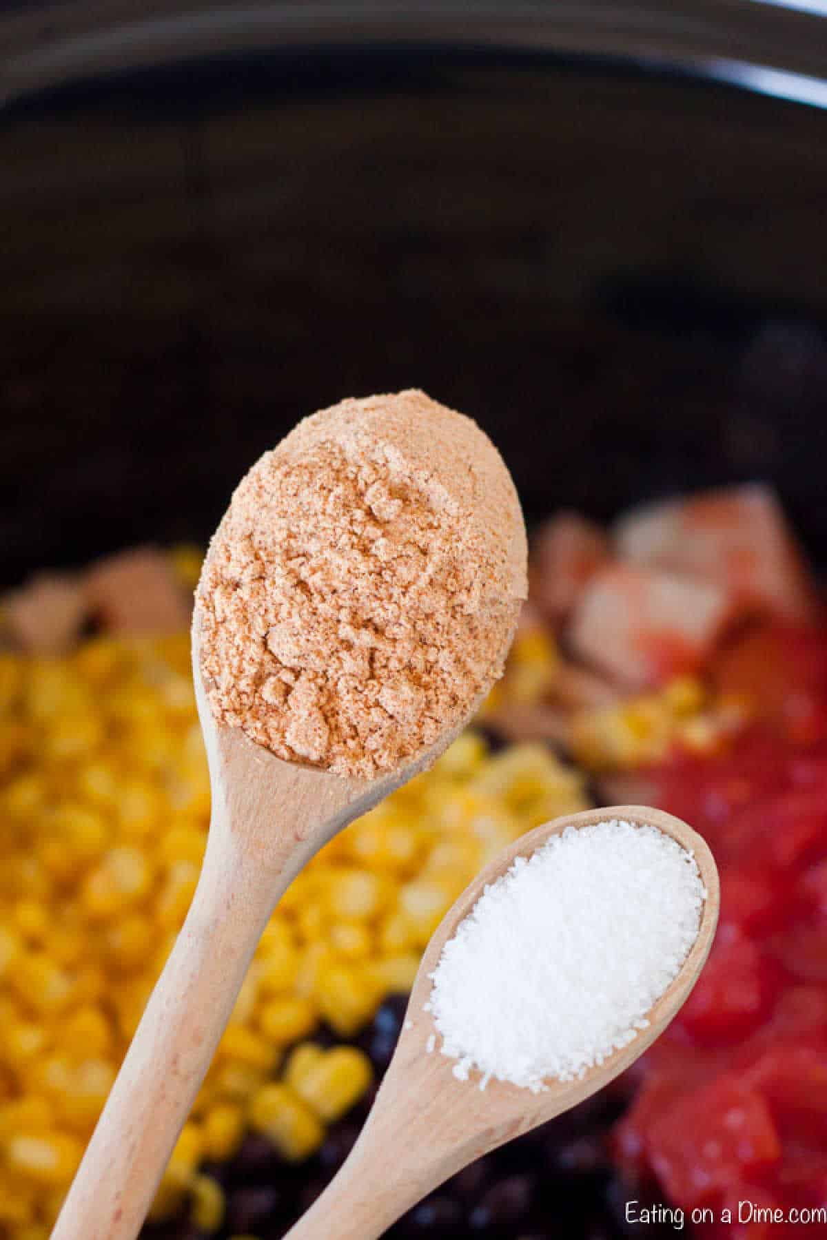 Close-up of two wooden spoons over a slow cooker for a delicious Slow Cooker Chicken Burrito Bowl Recipe. One holds a reddish-orange powder, the other, a white granular substance. Inside are black beans, corn, chopped tomatoes, and tender pieces of chicken.