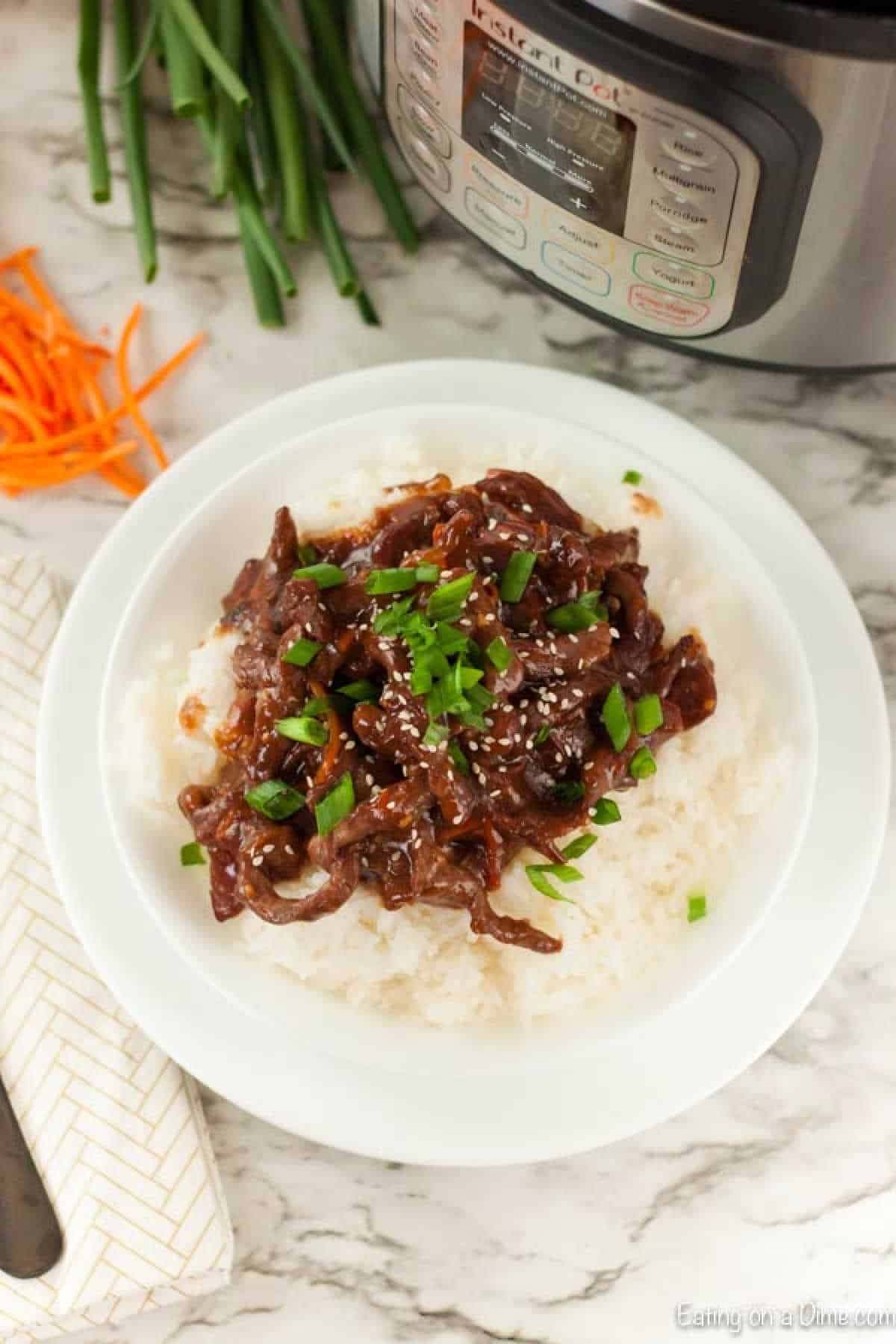 A plate of Instant Pot Mongolian Beef Recipe garnished with chopped green onions and sesame seeds, served over white rice. Chopped green onions and carrot slivers sit alongside the Instant Pot in the background on a marble surface.