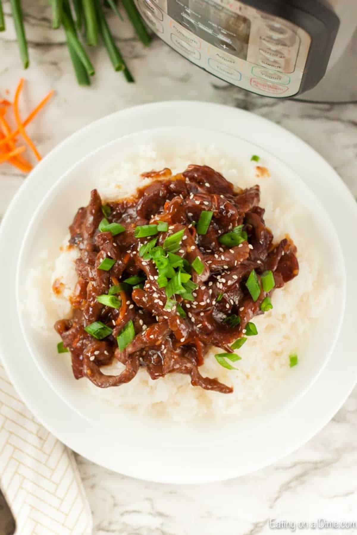 A plate of white rice topped with saucy Mongolian beef strips garnished with chopped green onions and sesame seeds. An Instant Pot and some sliced carrots are visible in the background, making it clear this is a delicious Instant Pot Mongolian Beef recipe.