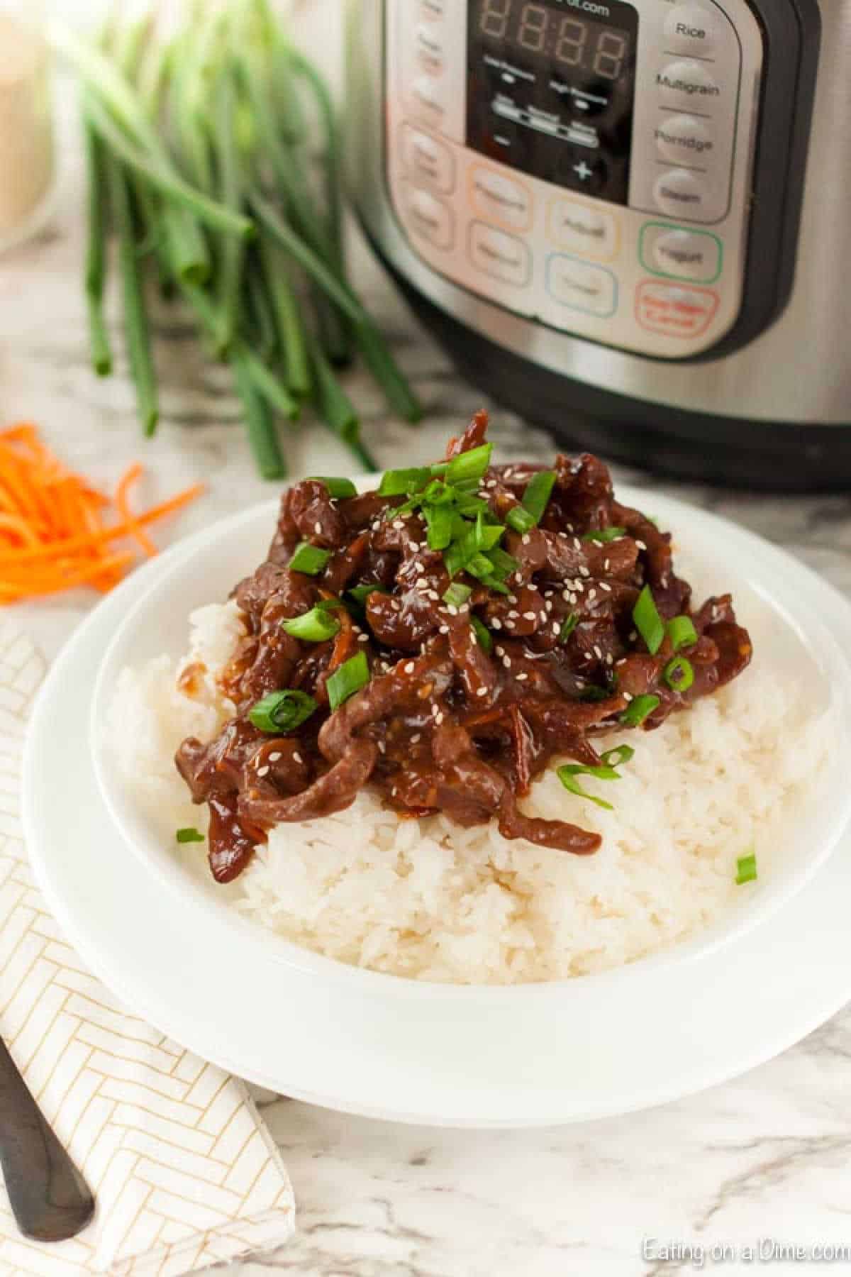 A plate of white rice is topped with savory Mongolian beef stir-fry, garnished with chopped green onions and sesame seeds. An Instant Pot and fresh green onions frame the scene on a marble countertop, hinting at this delicious recipe's quick and easy preparation.