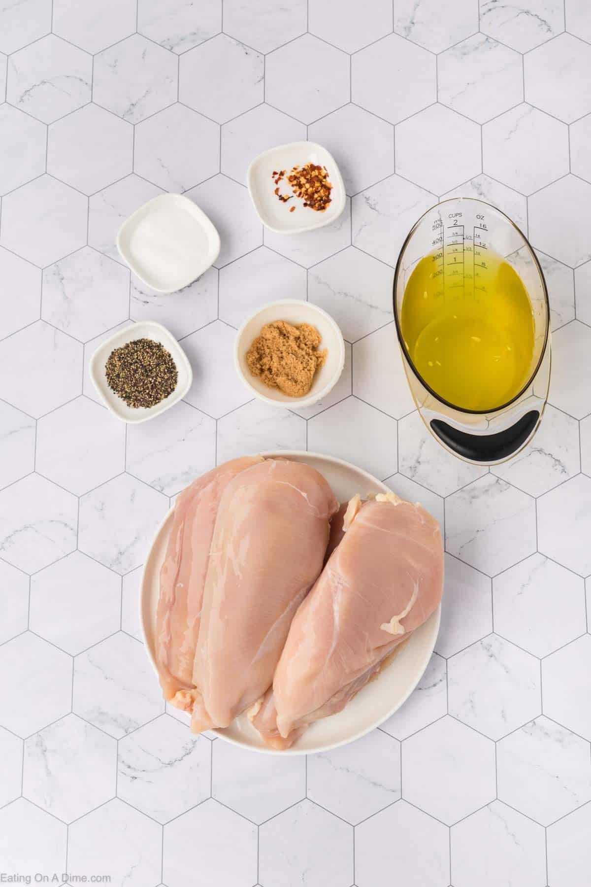 Raw chicken breasts on a plate, surrounded by small bowls of salt, black pepper, red pepper flakes, brown sugar, and a measuring cup with olive oil and pickle juice marinade. The background is a white hexagonal tile surface.