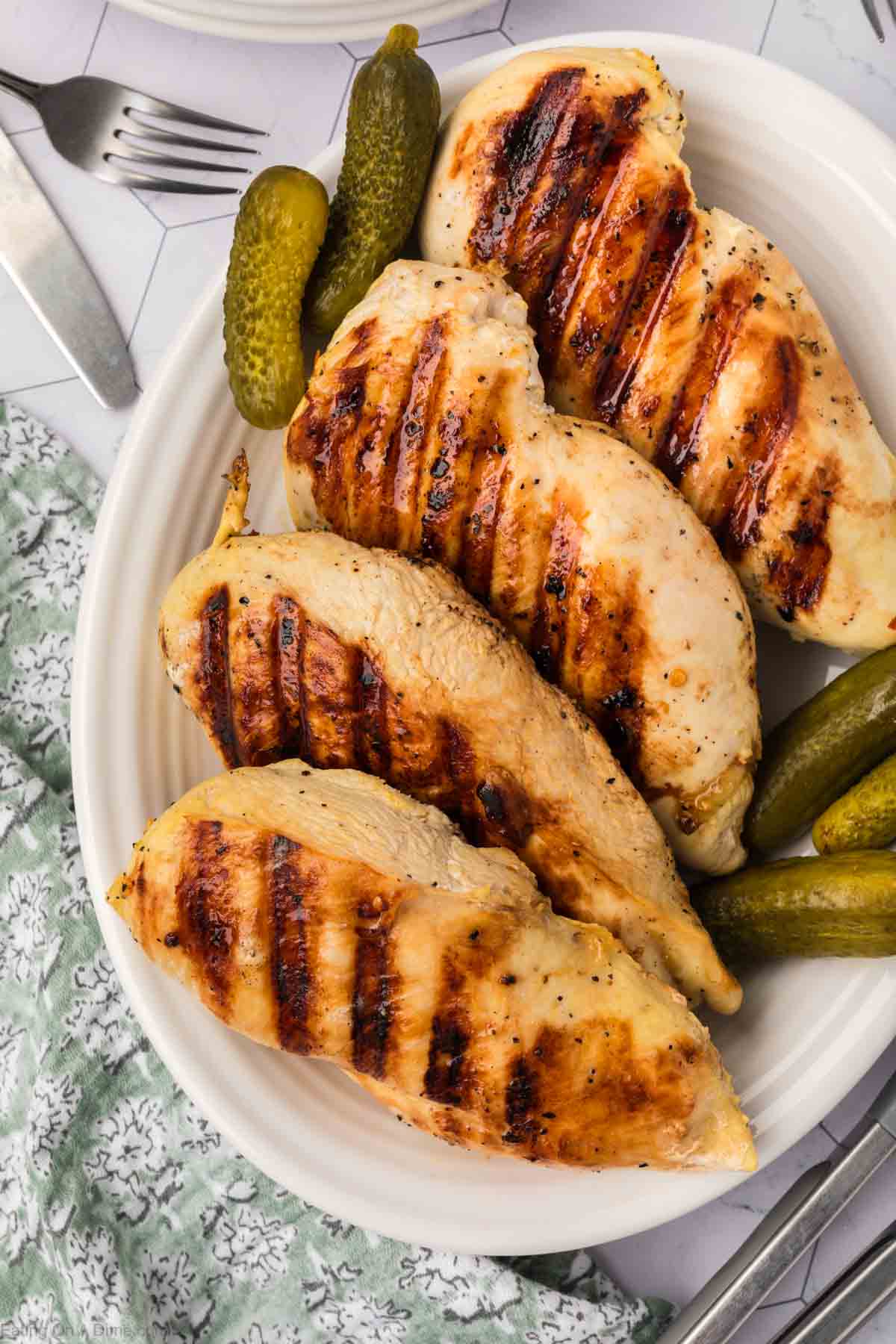 Grilled chicken breasts with char marks, infused with a tangy pickle juice marinade, sit on a white plate accompanied by crisp pickles. A floral cloth and cutlery subtly enhance the background.