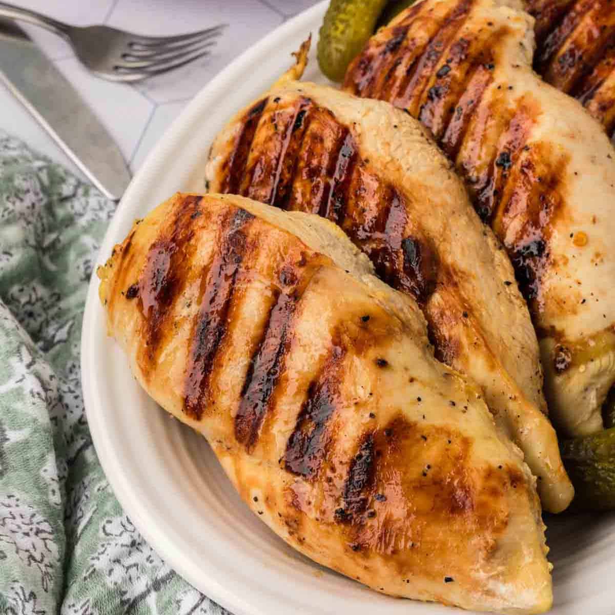 Grilled chicken breasts with prominent grill marks on a white plate, seasoned with a tangy pickle juice marinade and garnished with green pickles. A patterned green cloth and a fork and knife are placed to the side.