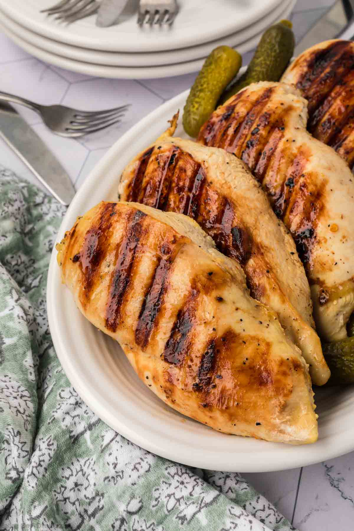 Grilled chicken breasts boast perfect grill marks, thanks to a tangy pickle juice marinade. They're served on a white plate with pickles against a patterned tablecloth, alongside utensils and a stack of white dishes.