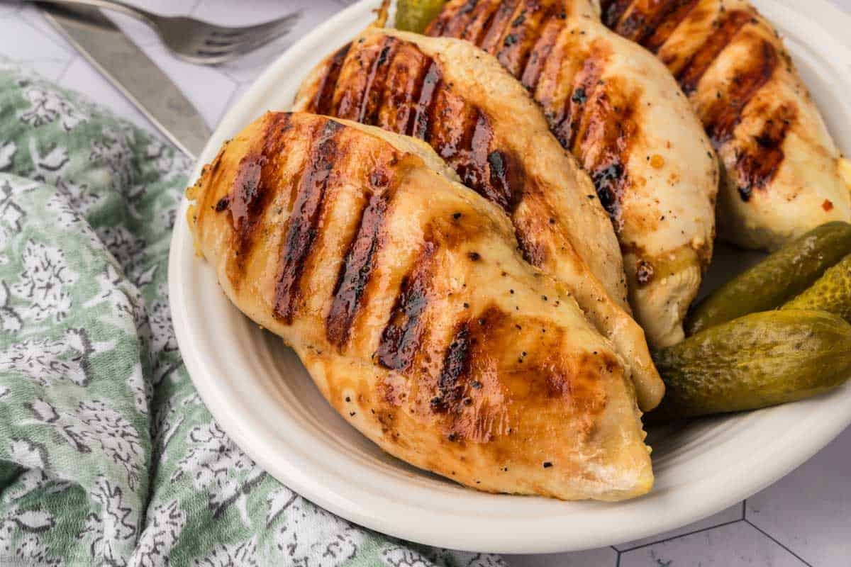 Grilled chicken breasts with char marks, marinated in tangy pickle juice, served on a white plate alongside crunchy pickles. A green patterned cloth adds a touch of color, with a fork partially visible in the background.