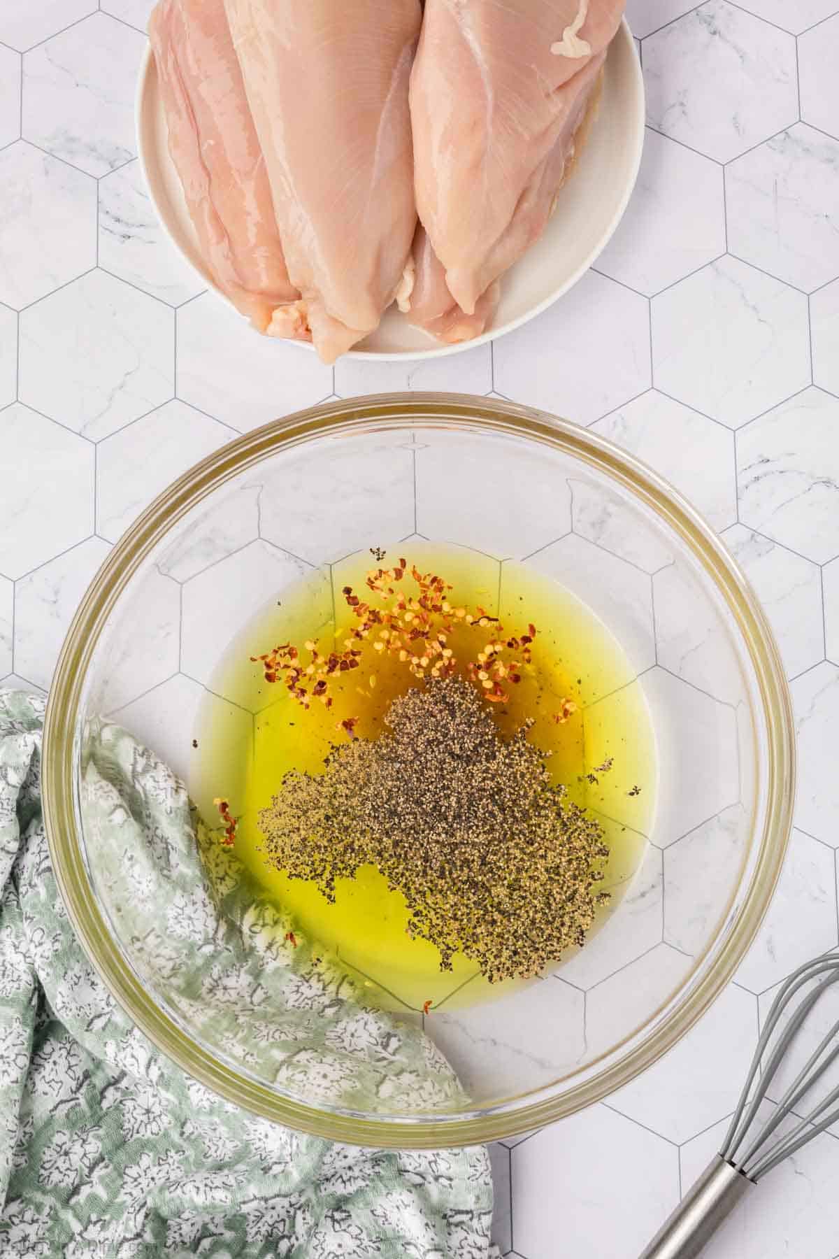 A glass bowl with olive oil, crushed red pepper, herbs, and a splash of pickle juice marinade sits on a hexagonal-patterned counter. Raw chicken breasts rest on a plate nearby. A green patterned cloth and a whisk are also positioned on the counter.