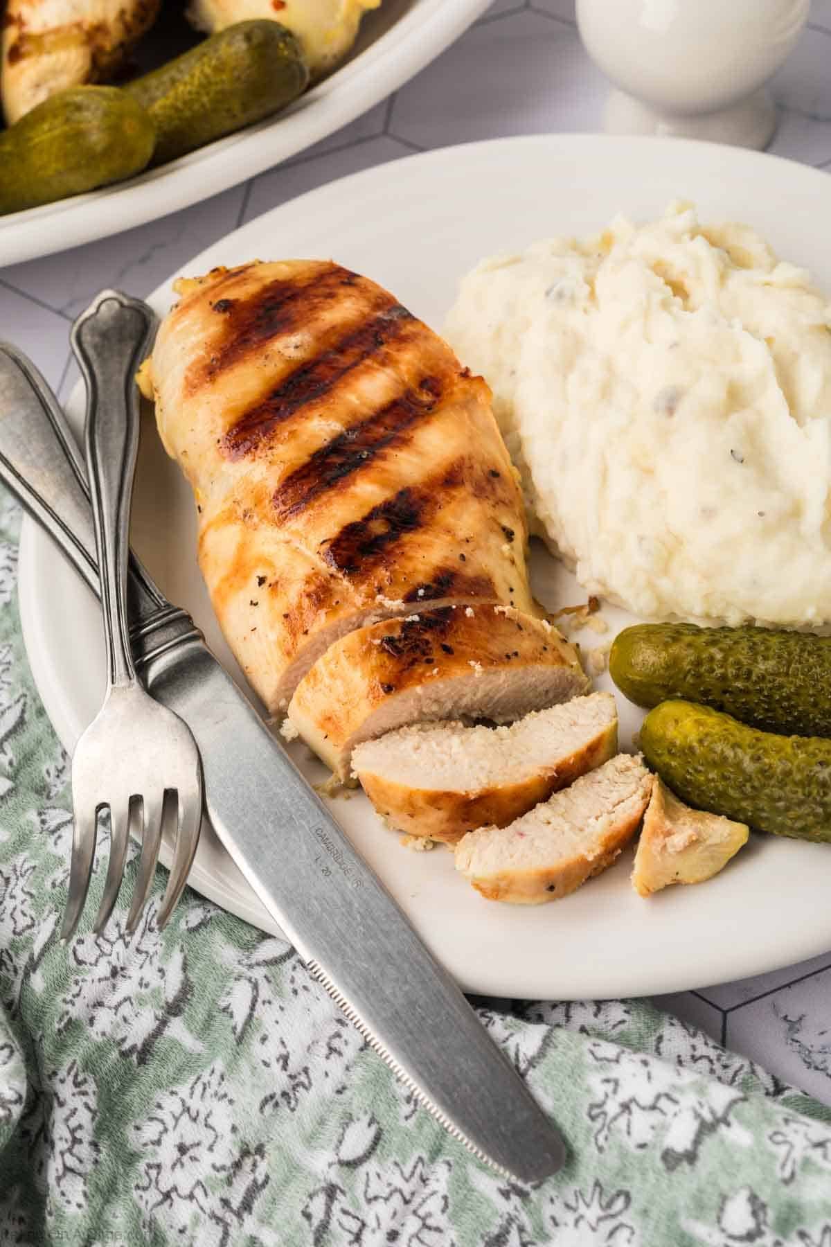 Grilled chicken breast, delicately infused with a pickle juice marinade, is slice-cut and served on a white plate accompanied by creamy mashed potatoes and tangy pickles. A fork and knife rest beside the dish on a green patterned cloth.