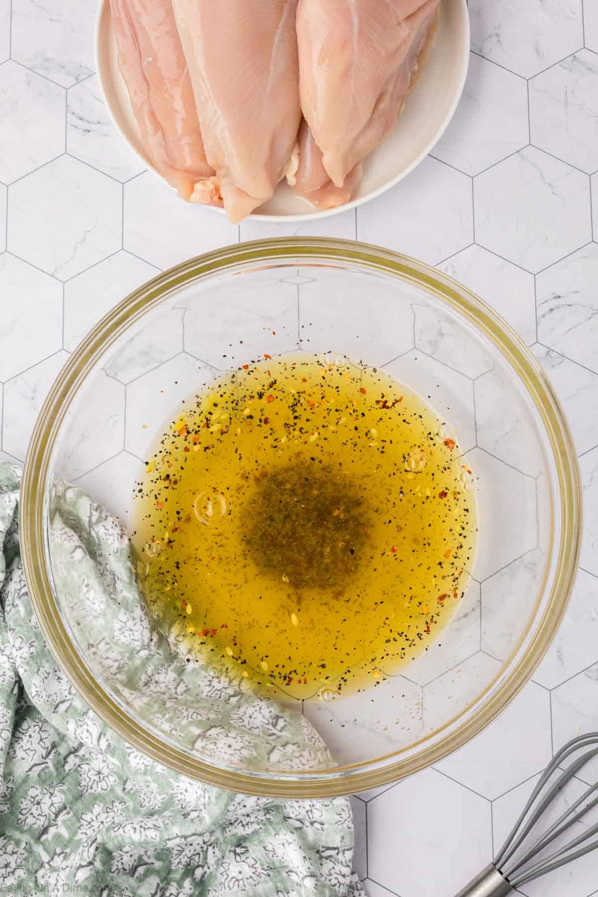 A glass bowl on a hexagonal patterned surface cradles a seasoned olive oil and pickle juice marinade, infused with herbs and spices. Nearby, a plate displays raw chicken breasts beside a green-patterned cloth, while a whisk rests ready for action next to the bowl.