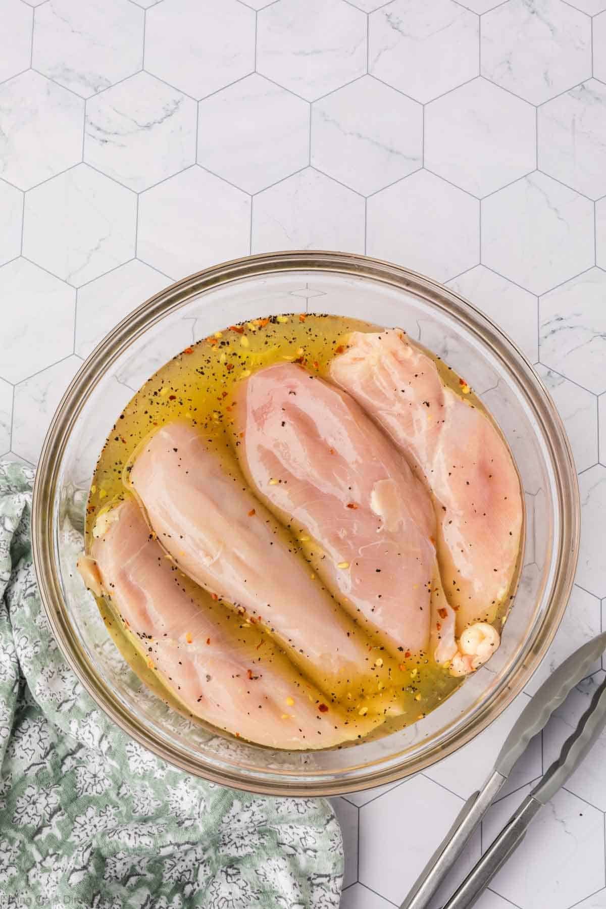 Four raw chicken breasts marinate in a bowl of seasoned oil with a hint of pickle juice. They're set on a hexagonal-patterned countertop, surrounded by a green floral cloth and shiny metal tongs.