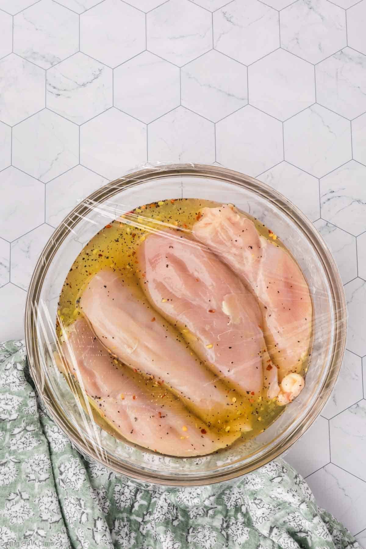 A glass bowl with raw chicken breasts soaking in a pickle juice marinade, covered with plastic wrap. The bowl rests on a hexagonal-tiled surface beside a green and white patterned cloth.