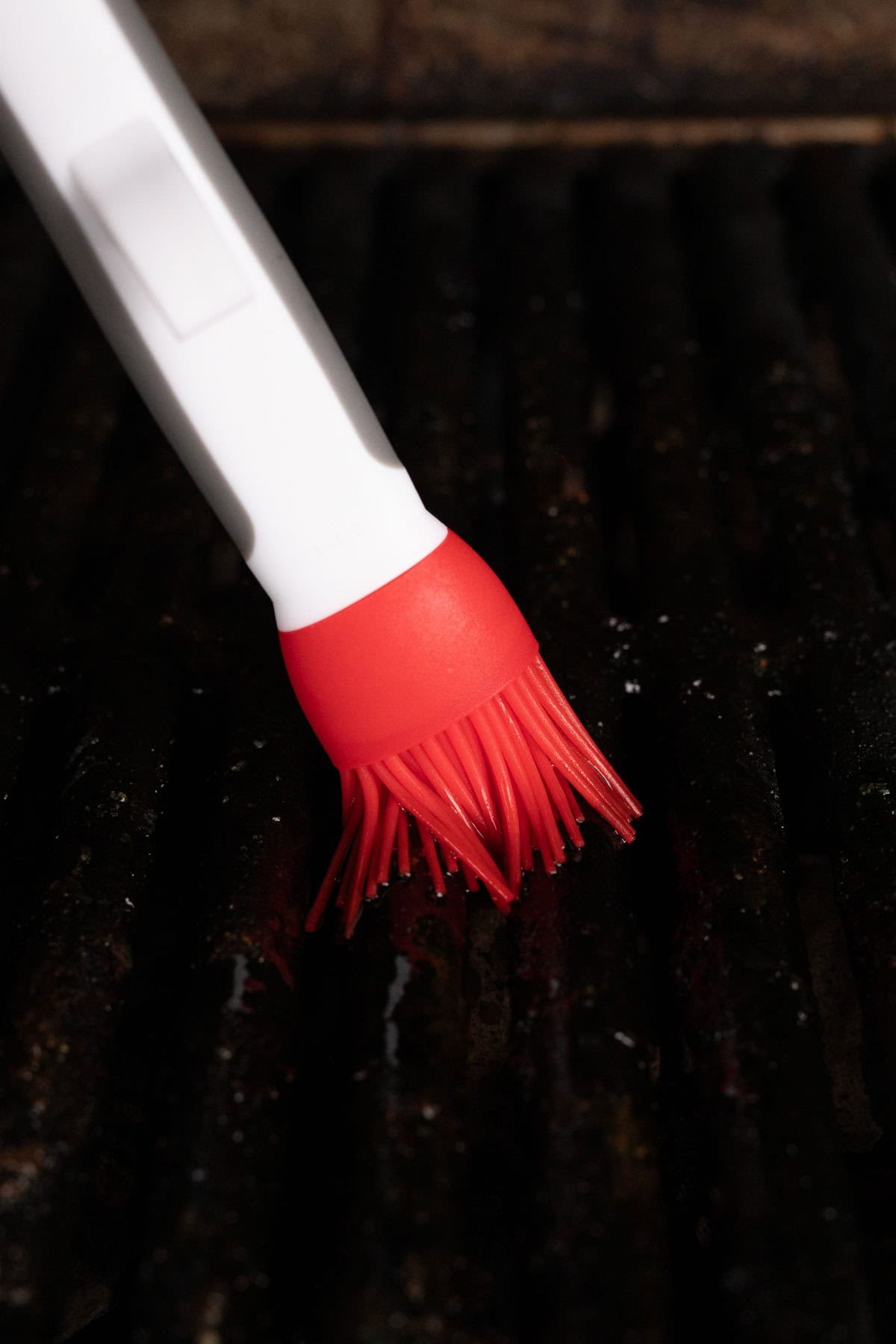 Close-up of a red silicone basting brush applying pickle juice marinade to a dark barbecue grill grate. The handle of the brush is white.