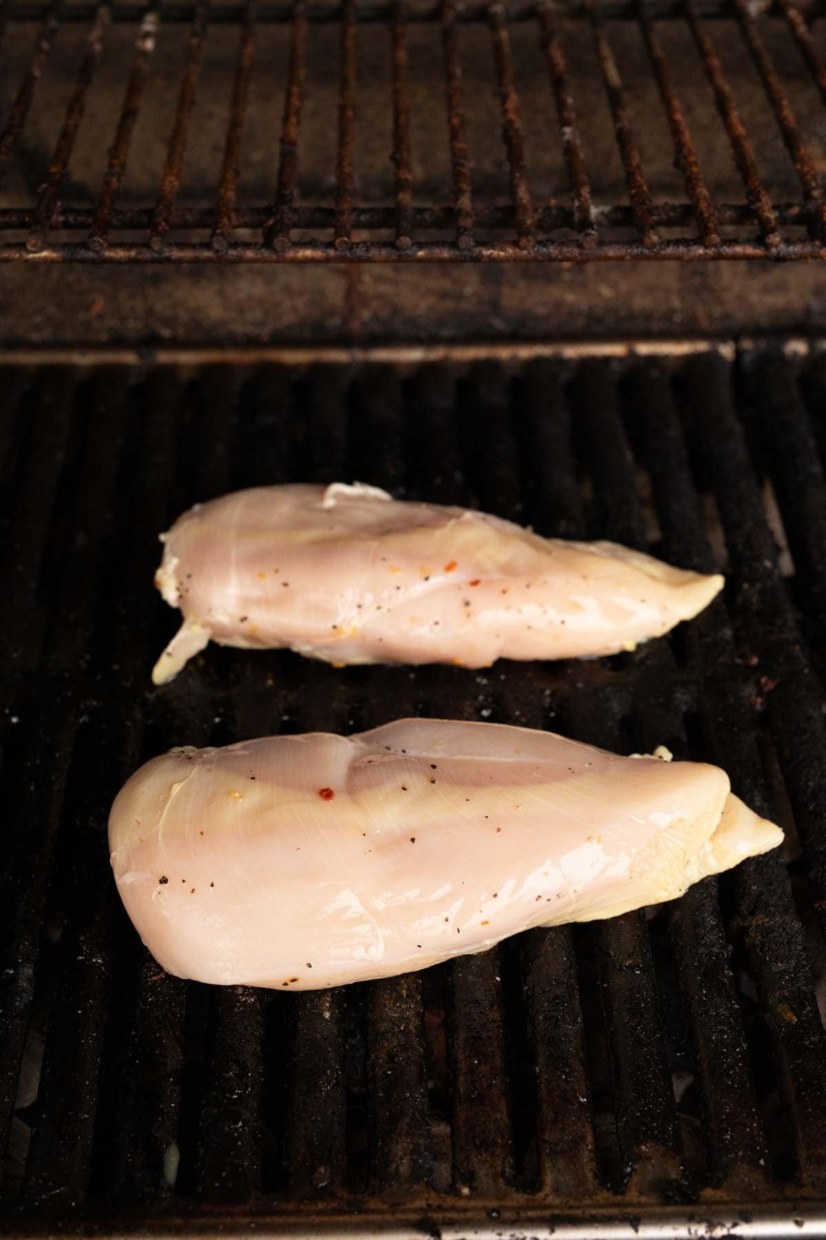 Two raw chicken breasts, marinated in pickle juice for a tangy twist, are placed on the well-used grill grates. Seasoned with a light sprinkle of black pepper and other spices, they’re ready to be cooked to perfection over the slightly charred bars.