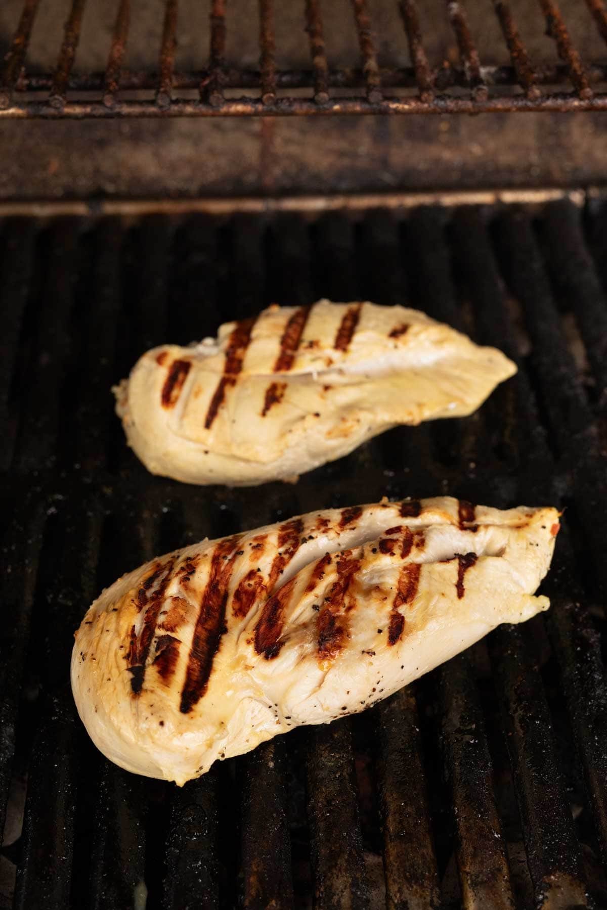 Two seasoned chicken breasts, marinated in pickle juice, sizzle on the barbecue, displaying bold grill marks. The meat is angled perfectly on the blackened grates, just beneath a partially visible upper shelf.