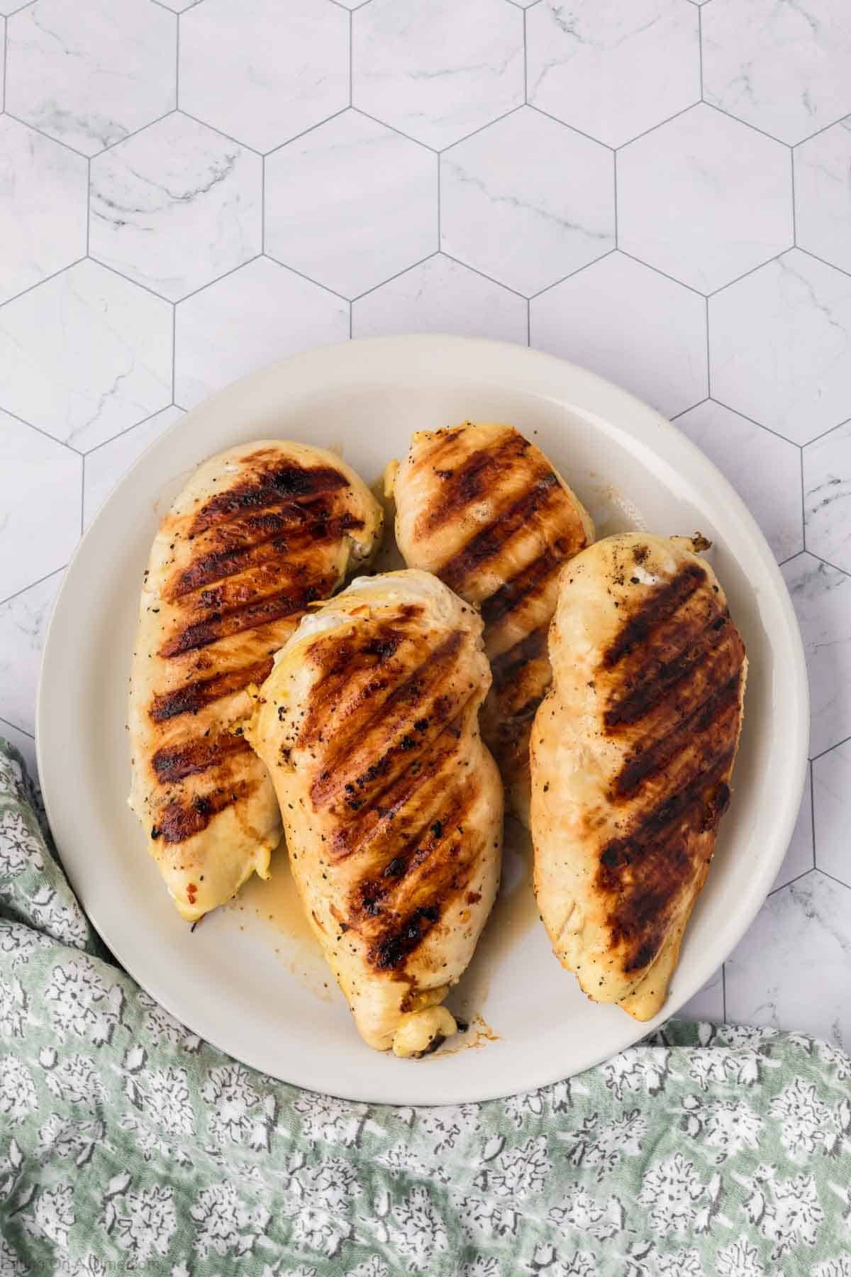 Grilled chicken breasts, marinated in tangy pickle juice, are arranged on a white plate. The golden-brown chicken boasts prominent grill marks. A green patterned cloth is partially visible beside the plate on a white, hexagonal-tiled surface.