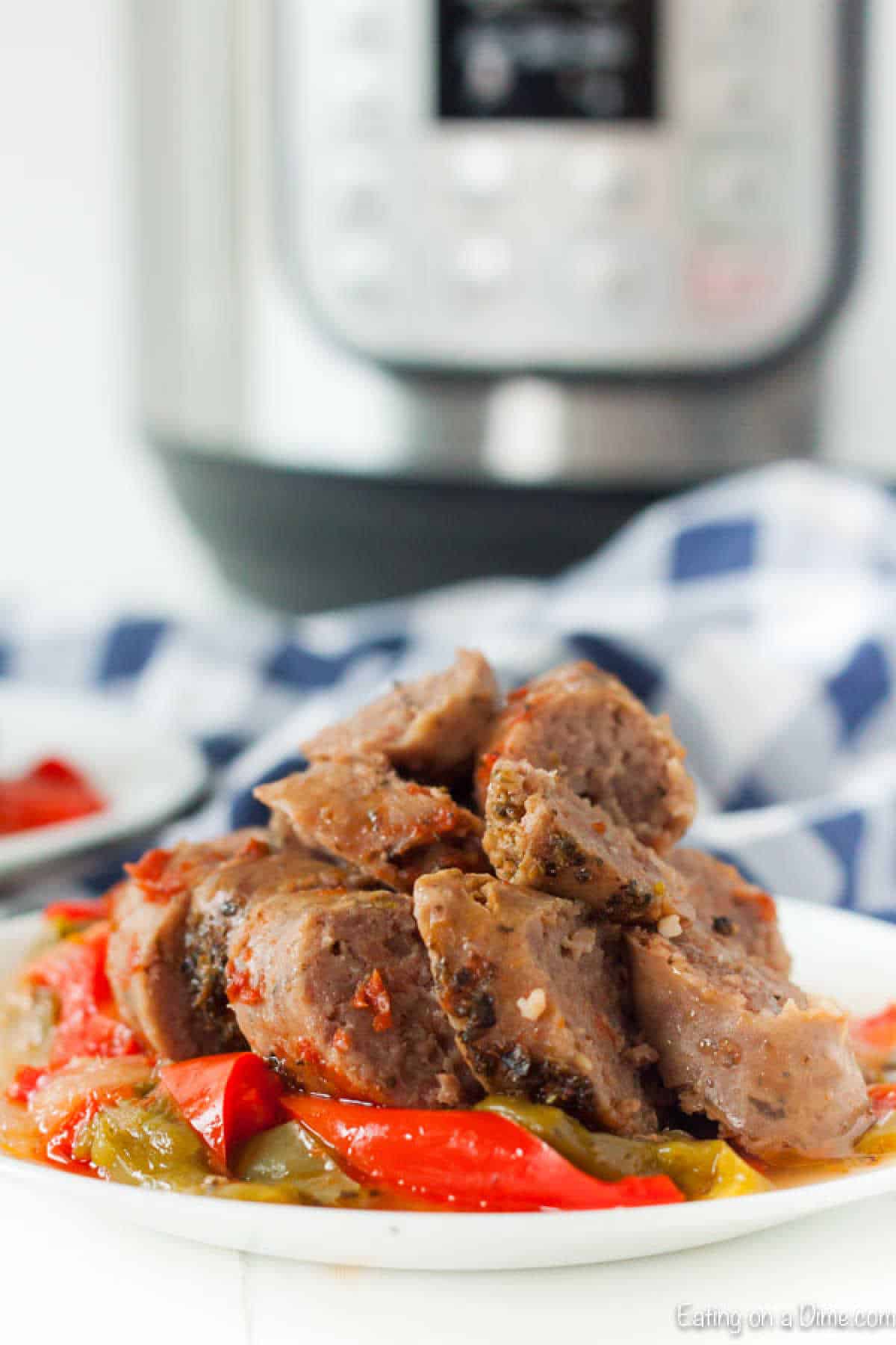 A plate of sliced sausage with red and green bell peppers sits enticingly in front of a blurred Instant Pot, promising a delicious Sausage and Peppers recipe. A blue and white checkered cloth adds charm to the background.