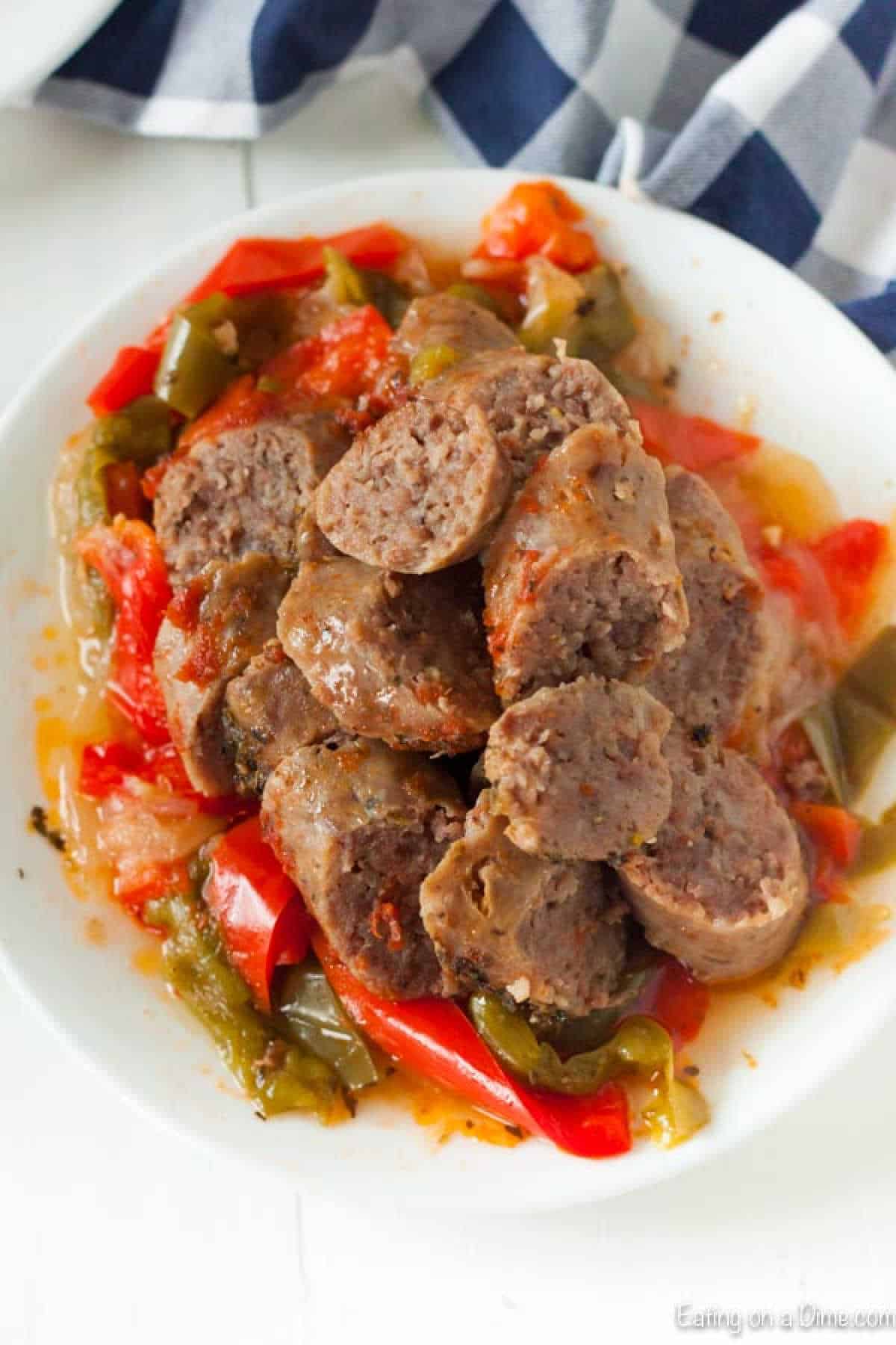 A plate of sliced sausages served over cooked red and green bell peppers in a white bowl on a light surface. A checkered cloth sits in the background, perfectly capturing the essence of an Instant Pot Sausage and Peppers recipe.