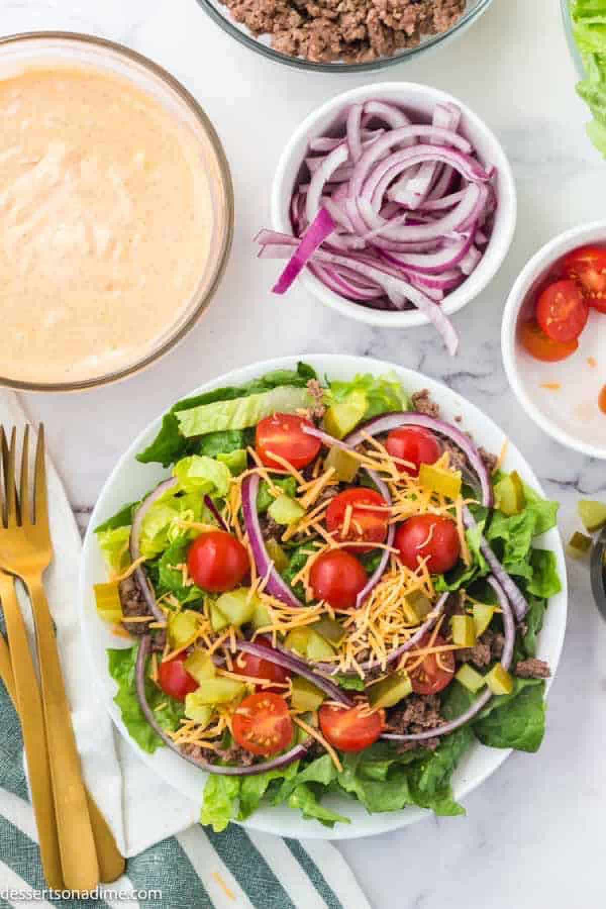 Bowl of big mac dressing, sliced red onions, cherry tomatoes, and a bowl of salad topped with red onions, cherry tomatoes and shredded cheese with ground hamburger meat