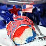 A slice of red, white and blue cake adorned with a drizzle of red icing, a small American flag, and shiny star toppers sits invitingly on a white plate. A silver fork lies beside it, poised against a backdrop of star-patterned fabric.