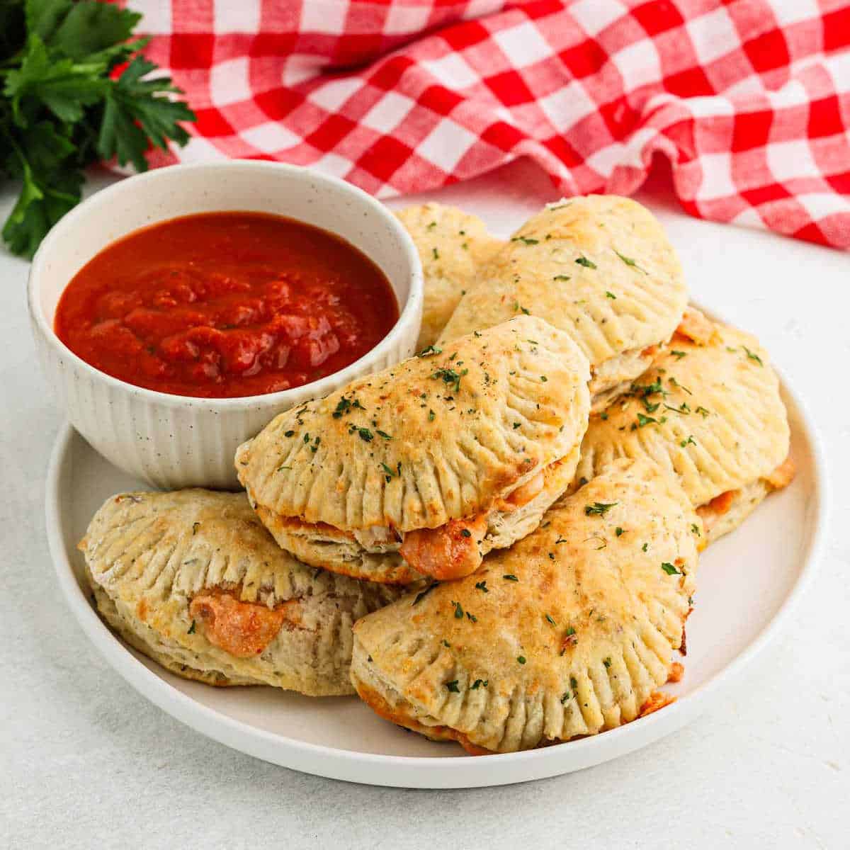 A plate of baked empanadas filled with cheese and herbs, reminiscent of a classic pizza pocket recipe, is garnished with parsley. A bowl of tomato sauce sits beside them on a white surface, with a red and white checkered cloth in the background.