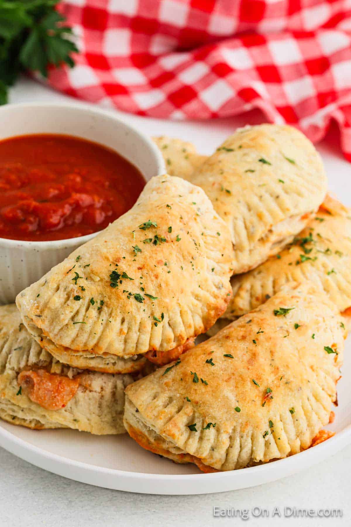 A plate of golden-brown empanadas, reminiscent of a classic pizza pocket recipe, is arranged around a bowl filled with red marinara sauce. A red and white checkered cloth is partially visible in the background.