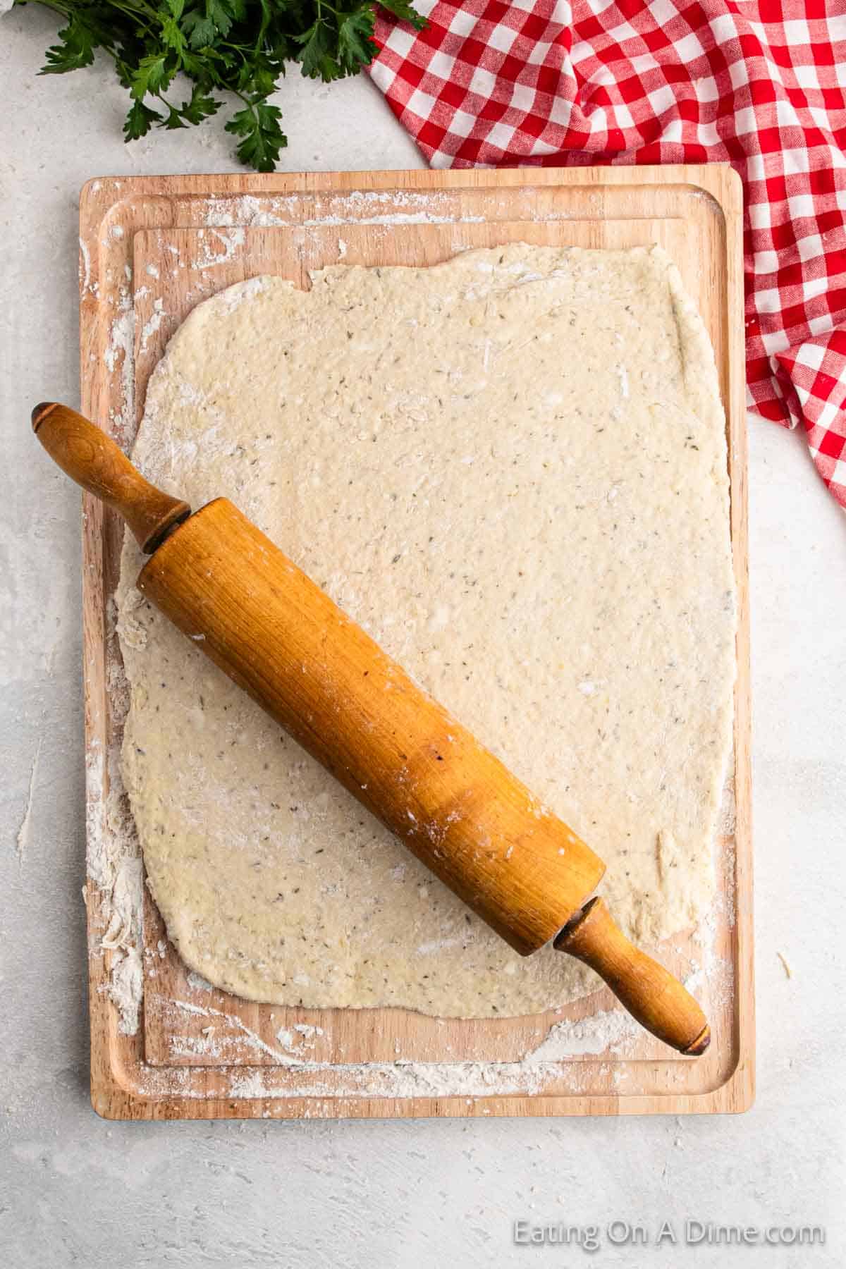 A wooden rolling pin rests on a sheet of flattened dough, perfect for crafting a pizza pocket recipe, placed on a wooden cutting board. A red and white checkered cloth is partly visible in the corner, with parsley leaves nearby.