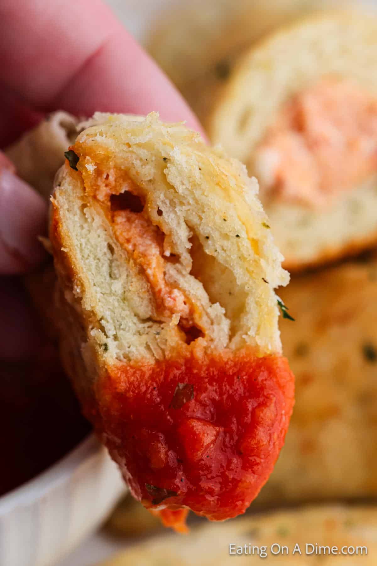 A close-up of a hand holding a pizza pocket brimming with melted cheese and herbs, with tomato sauce at one end. More pieces are blurred in the background.