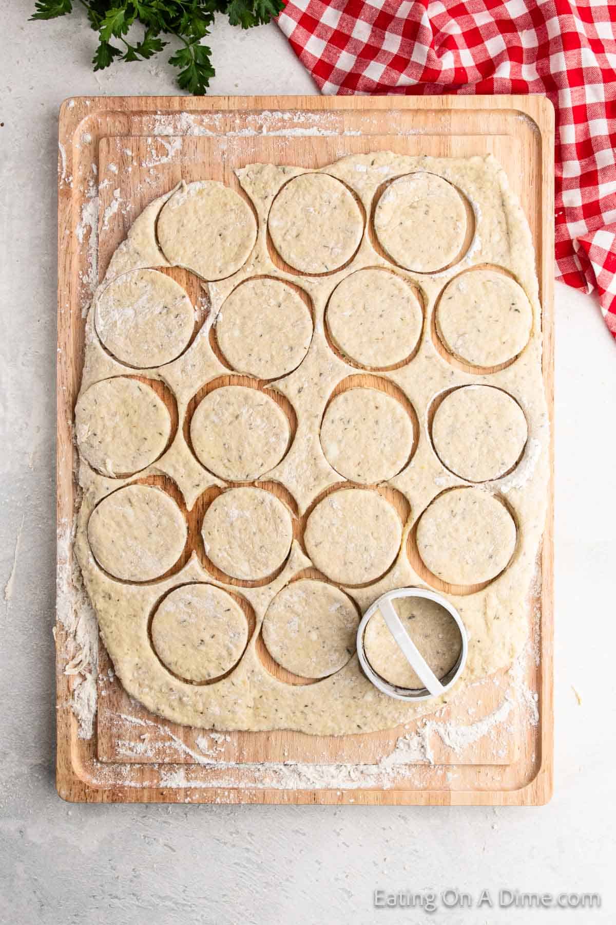 A wooden board displays rolled-out dough circles, crafted with a round cookie cutter for a delicious pizza pocket recipe. Fresh herb specks adorn the dough, while a red and white checkered cloth and sprigs of parsley add charm to the background.