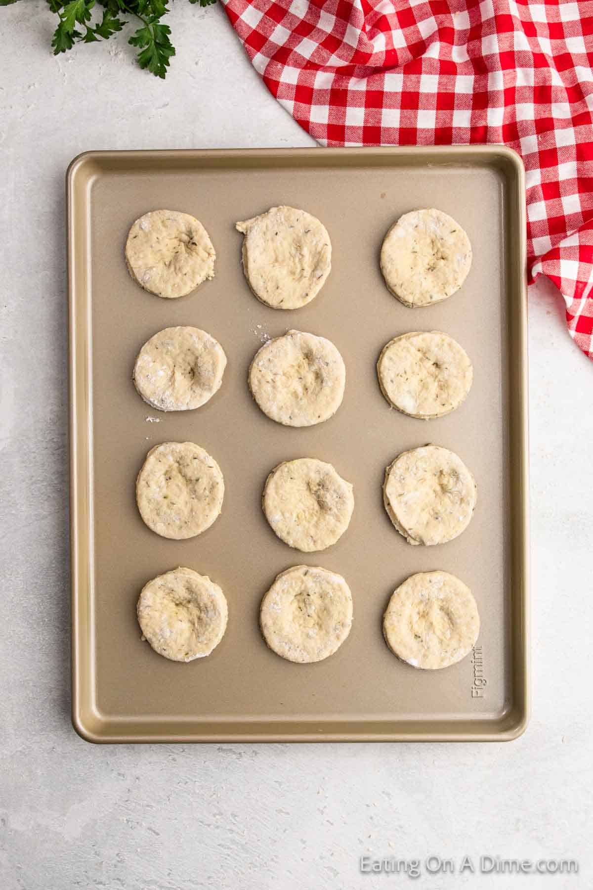 A baking tray holds twelve evenly spaced unbaked biscuit dough rounds, perfect for a pizza pocket recipe. A red and white checkered cloth peeks from the top right corner, while a sprig of parsley rests in the top left on a light-colored surface.