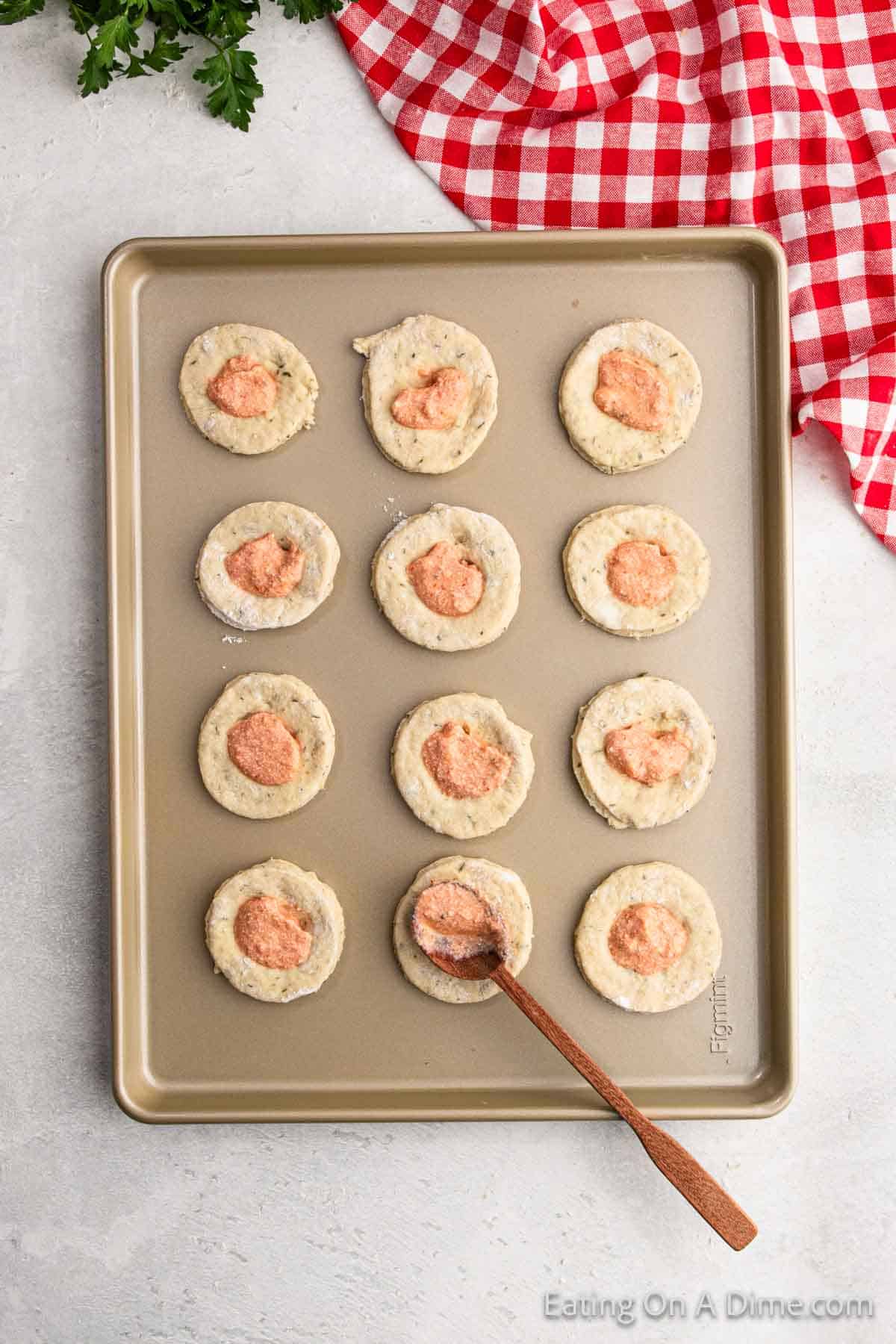 A baking tray holds twelve uncooked savory dough rounds, the essence of a pizza pocket recipe, each with a dollop of reddish filling. A wooden spoon gracefully rests on one round, while a red and white checkered cloth adds charm in the background.