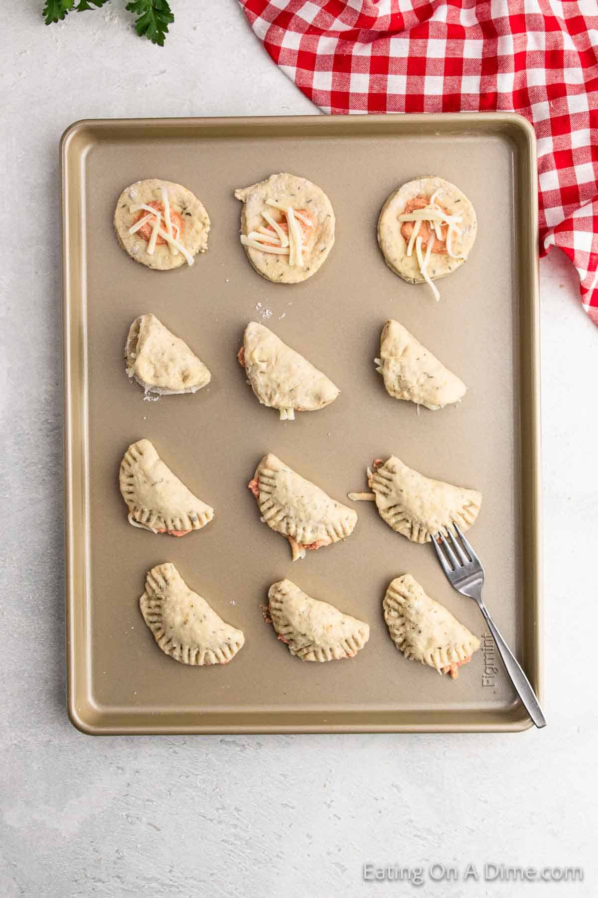 A baking tray on the countertop with puff pastry rounds awaits transformation into a Pizza Pocket recipe. Some are filled with shredded cheese and meat, folded into half-moons, and sealed with a fork. A red and white checkered cloth is nearby, adding a touch of charm.