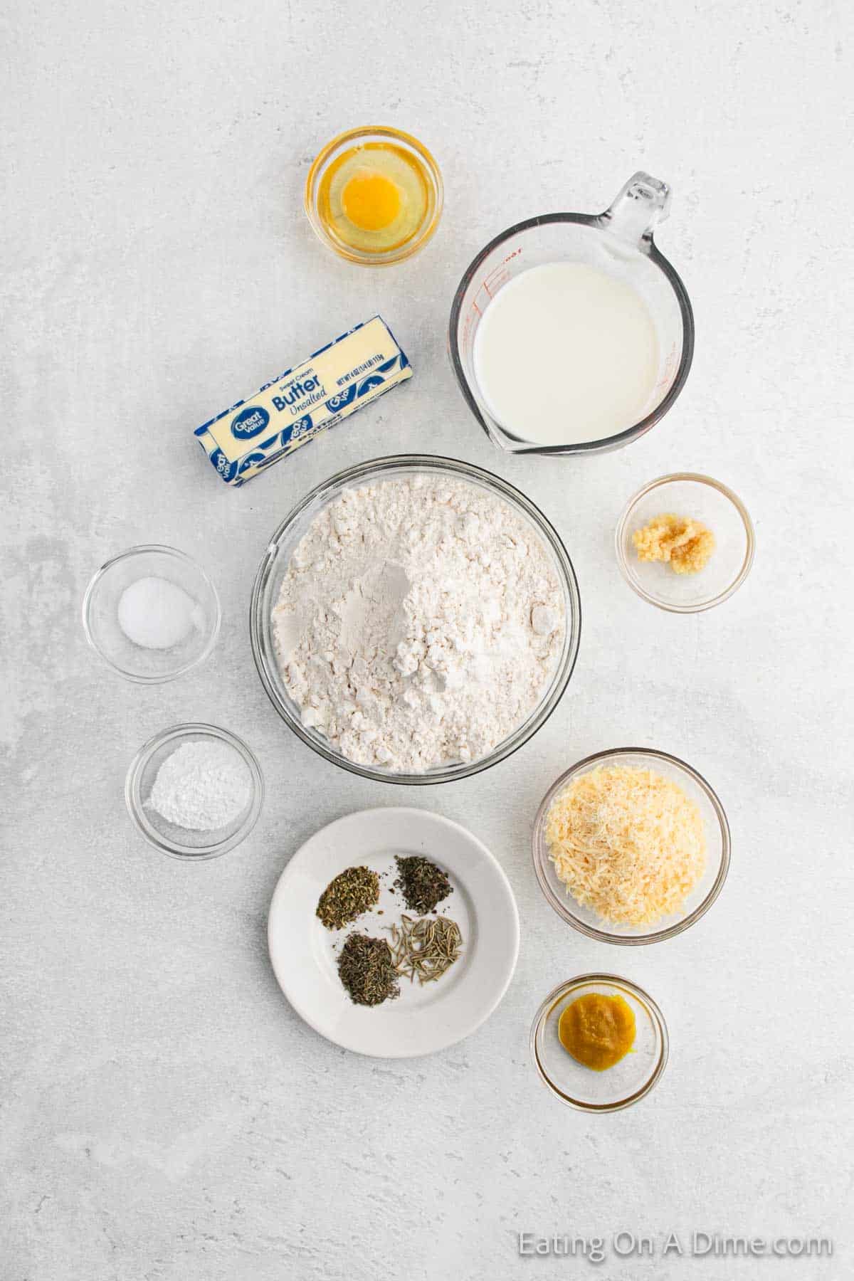 Various baking ingredients are arranged on a light surface: a bowl of flour, a stick of butter, grated cheese, a jug of milk, and small bowls containing salt, sugar, herbs, garlic paste, and an egg yolk—all set for crafting the perfect pizza pocket recipe.