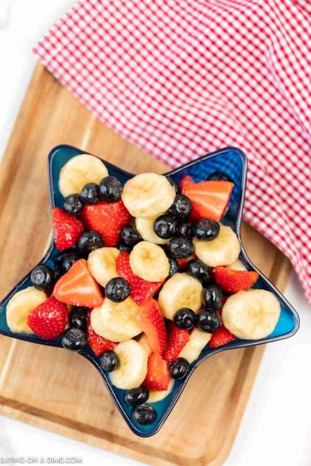 A star-shaped blue bowl brimming with a red, white, and blue fruit salad showcases sliced bananas, strawberries, and blueberries. The vibrant bowl rests on a wooden surface beside a red and white checkered cloth.