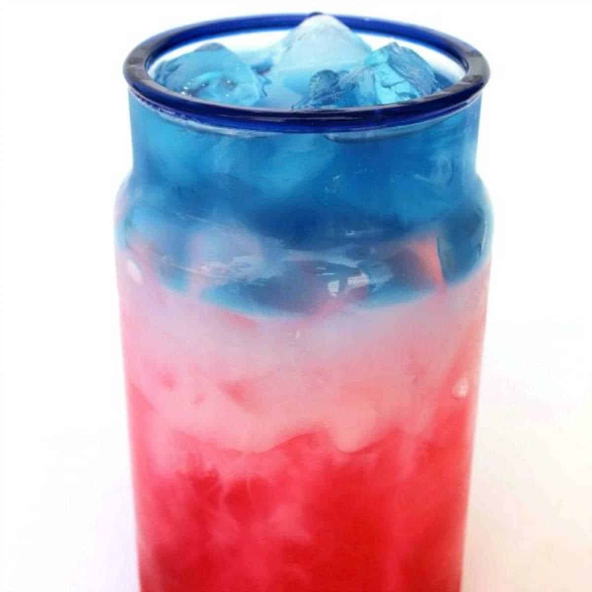 A red, white, and blue drink elegantly layered in a transparent glass with a striking blue rim. The vibrant beverage features icy layers of blue at the top, white in the middle, and red at the base, offering a refreshing visual delight.