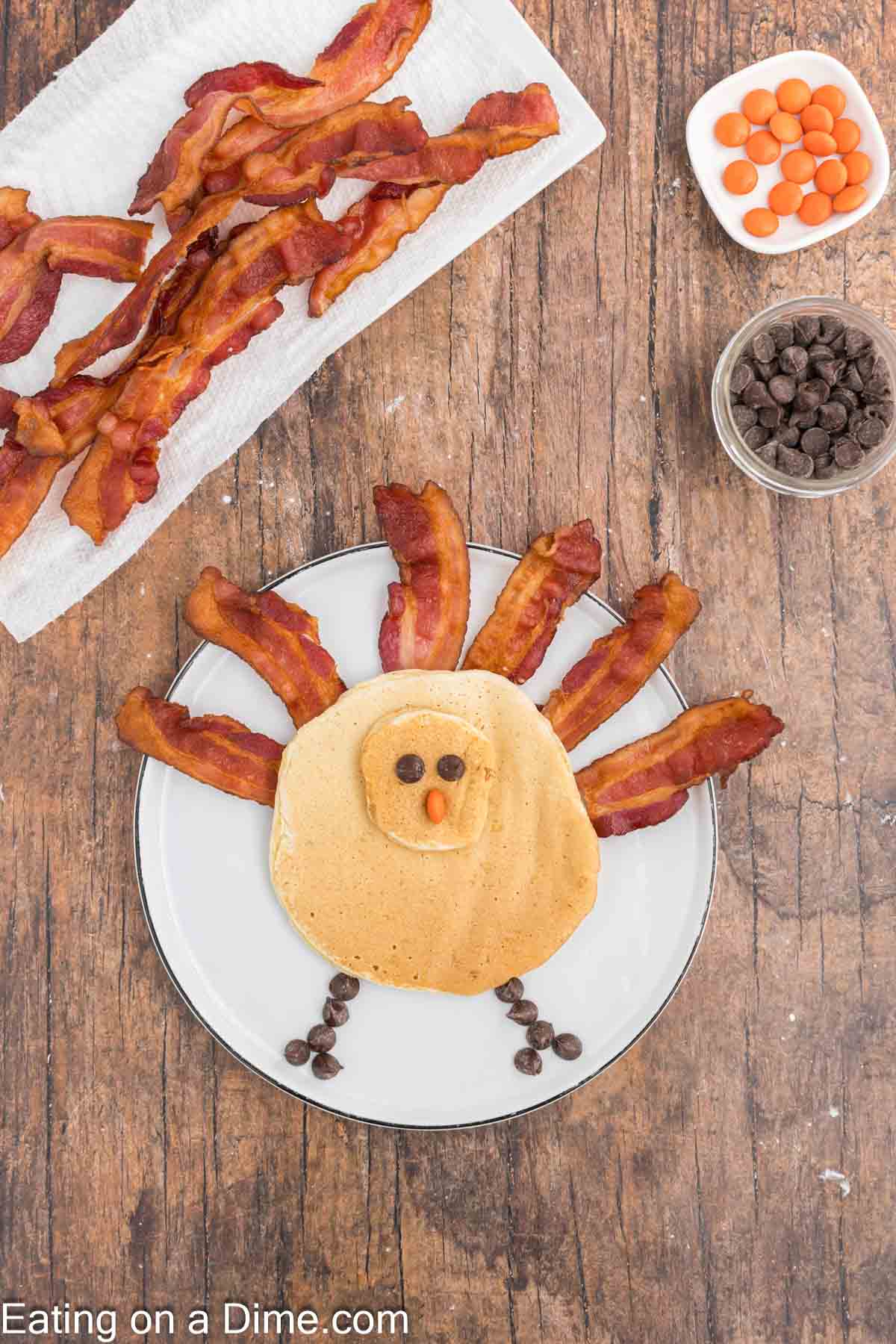 A breakfast plate featuring a turkey pancake, cleverly crafted with bacon strips as feathers, chocolate chips for eyes and feet, and an orange candy for the beak. It's surrounded by extra bacon on a paper towel and bowls of chocolate chips and orange candies.