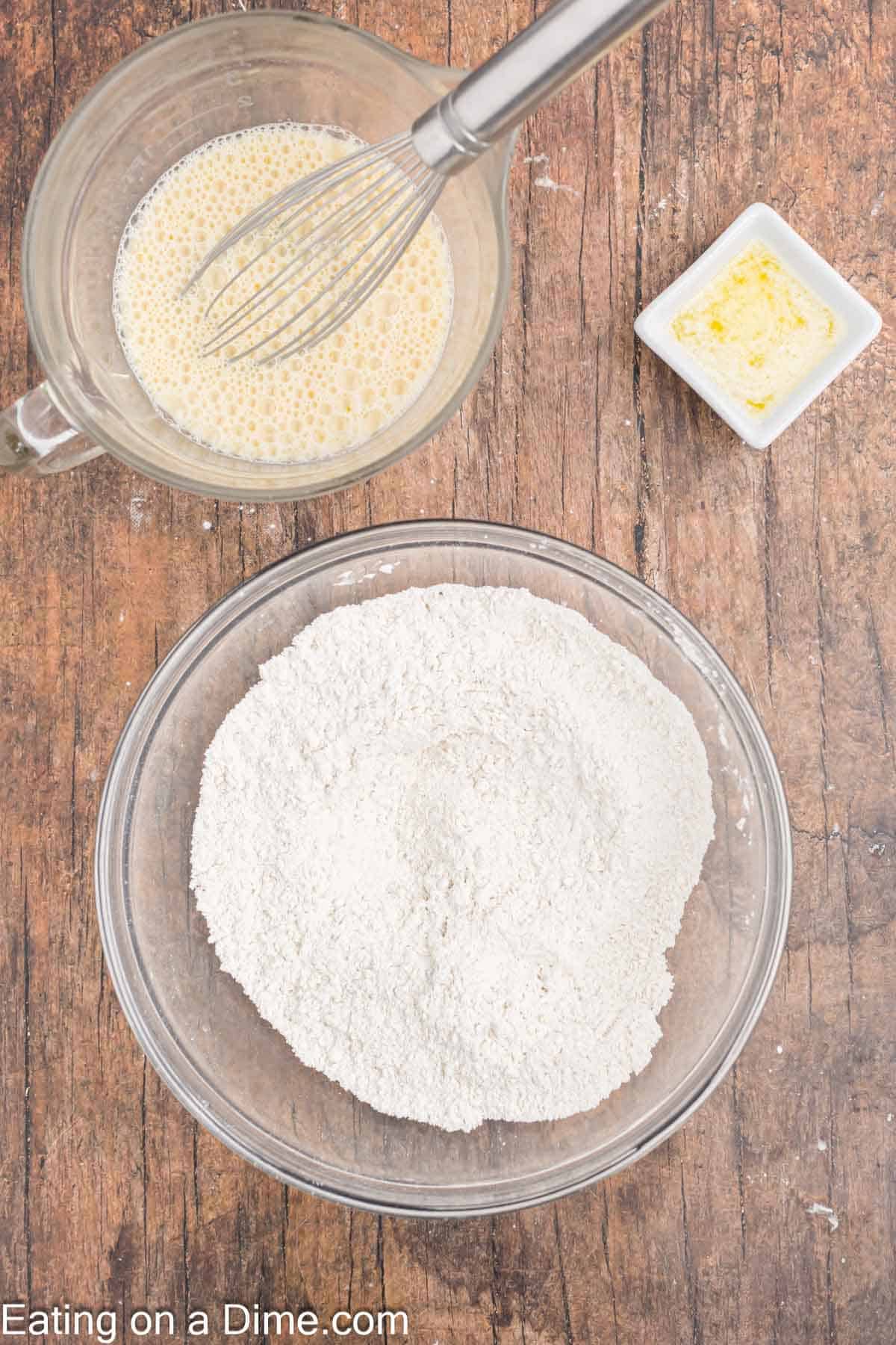 A top-down view of a wooden table with baking ingredients for turkey pancakes. A bowl of flour sits below a measuring cup filled with a whisked egg mixture. A small dish of melted butter is nearby, ready to add richness to the savory breakfast delight.