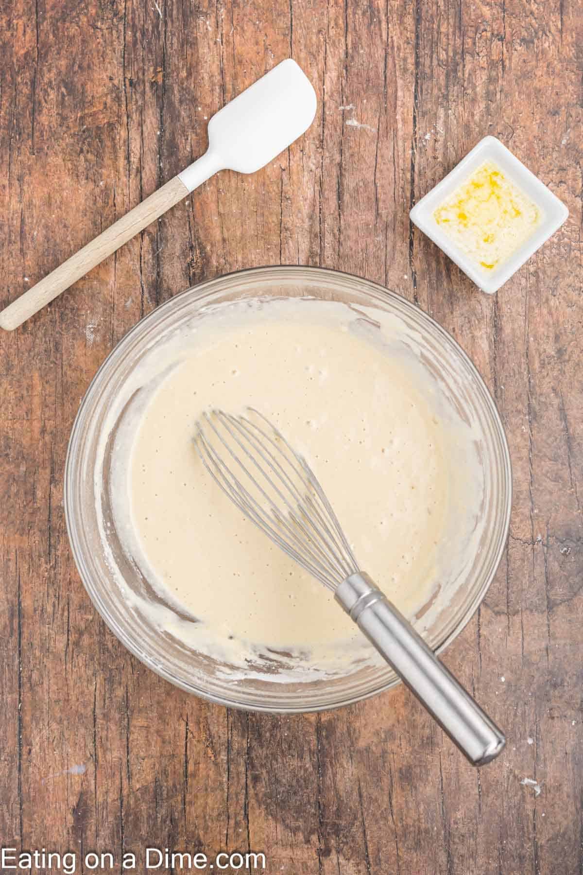 A glass bowl with pancake batter, perfect for crafting delightful turkey pancakes, and a whisk sits on a wooden table. A spatula rests nearby, while a butter dish occupies the corner. Text at the bottom reads "Eating on a Dime.com".