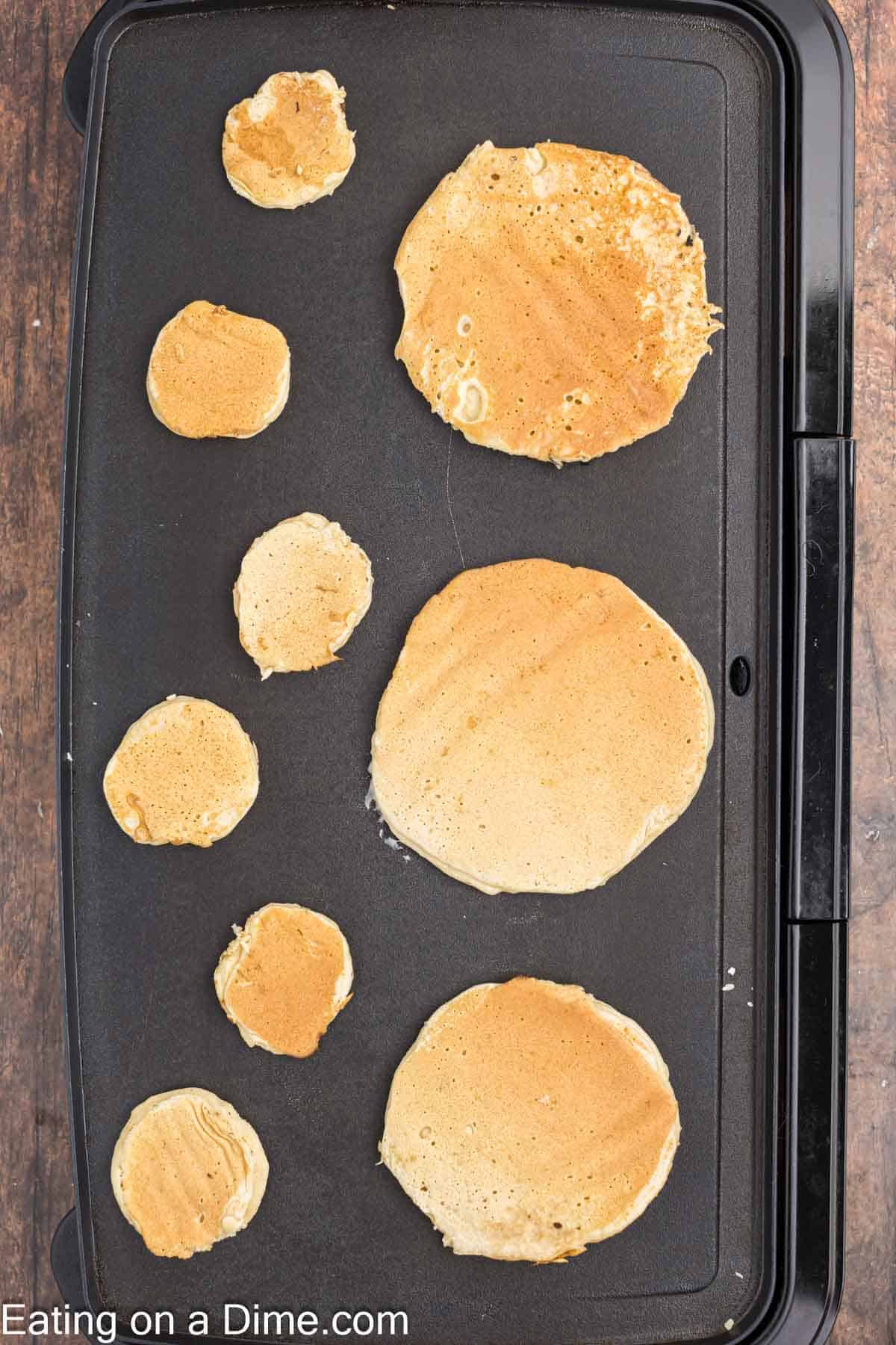 A variety of golden-brown turkey pancakes sizzle on the griddle. The sizes, ranging from small to large, are irregularly placed over the black cooking surface. The text "Eating on a Dime" is visible in the bottom left corner.