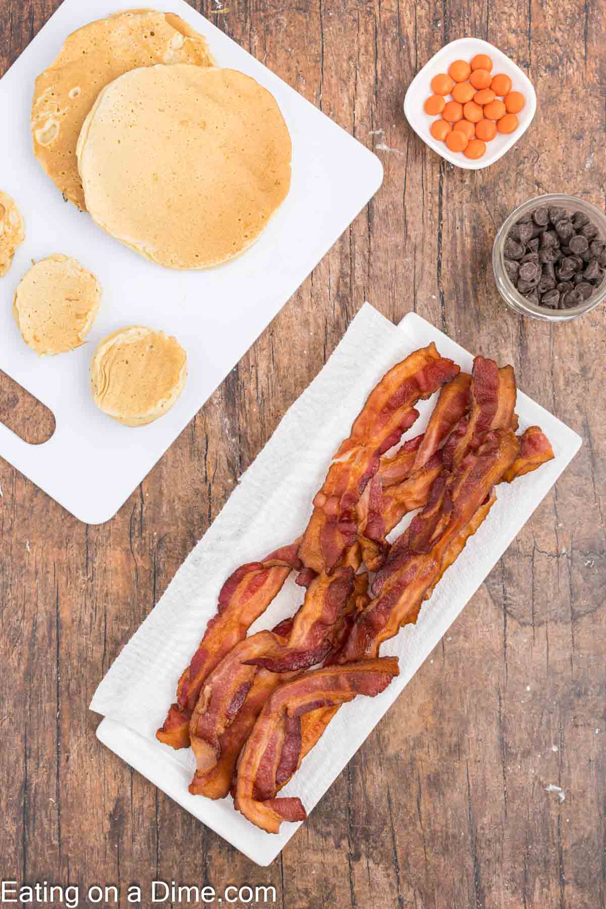 A wooden table showcases a delightful feast featuring turkey pancakes on a white cutting board, crispy bacon resting on a paper towel, and small bowls filled with orange candies and chocolate chips nearby.