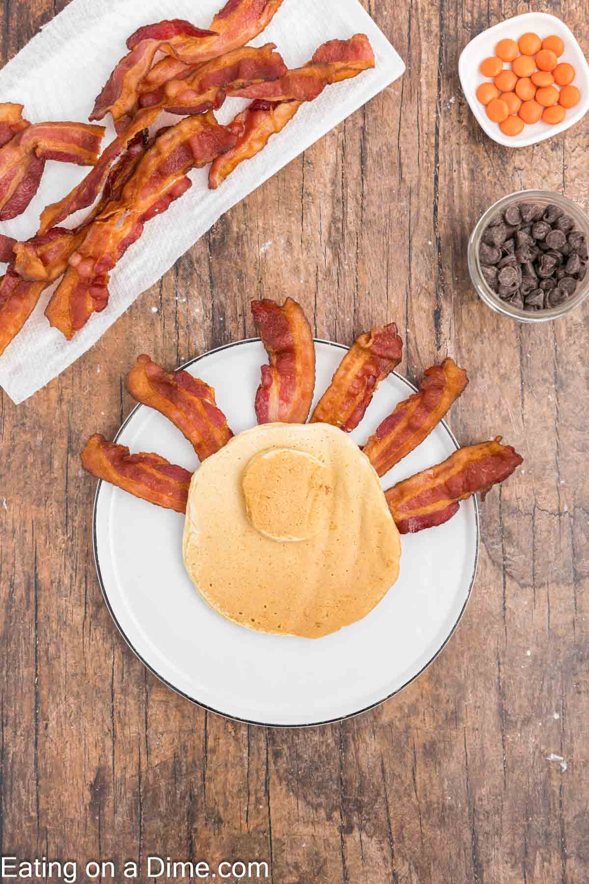 A turkey pancake is artfully designed to resemble a turkey on a white plate, with strips of bacon cleverly arranged as tail feathers. In the background, more bacon rests on a paper towel alongside small bowls of chocolate chips and candy on the wooden table surface.