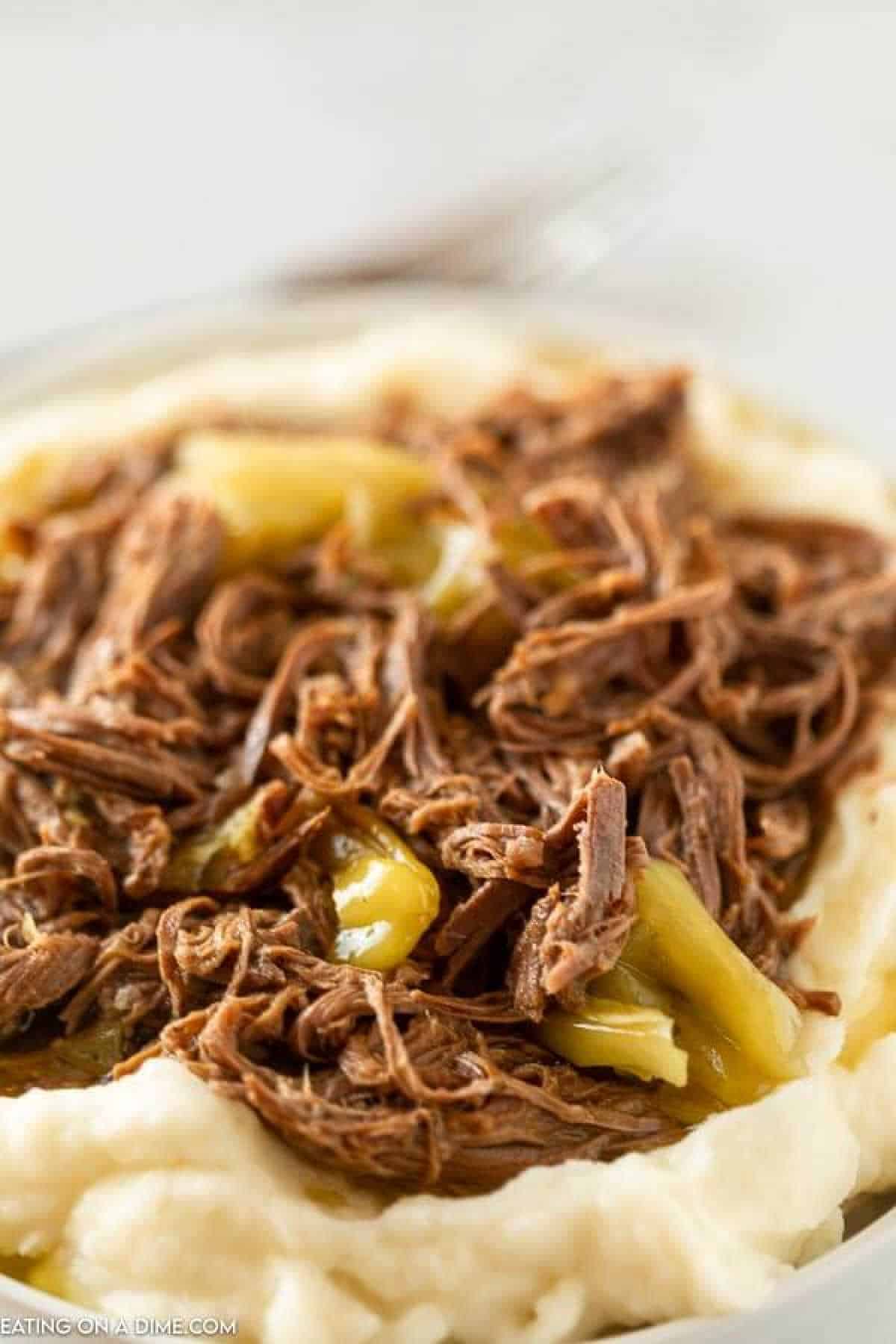 A plate of creamy mashed potatoes topped with Mississippi Pot Roast, featuring tender shredded beef and sliced green peppers. The dish appears savory and appetizing, with the roast beef and peppers adding texture and color to the smooth mashed potatoes.