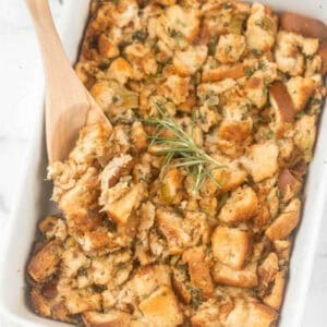 A white baking dish filled with golden-brown bread stuffing mixed with herbs and chopped celery. A wooden spoon is scooping a portion of the stuffing from the dish. Made with the best bread for stuffing, a sprig of fresh rosemary is placed on top as garnish.