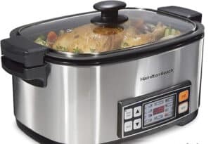 A stainless steel slow cooker, known as one of the best multicookers, features a digital display showing a cooked whole chicken with vegetables. Through the glass lid, steam reveals carrots and herbs. The control panel offers intuitive buttons and a clear time display for effortless cooking.