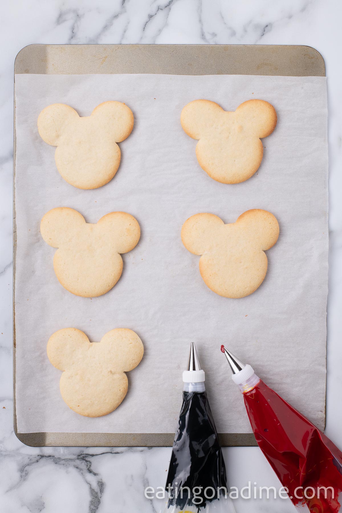 Baked mickey mouse cookies on a baking sheet with black icing and red icing in a piping bag